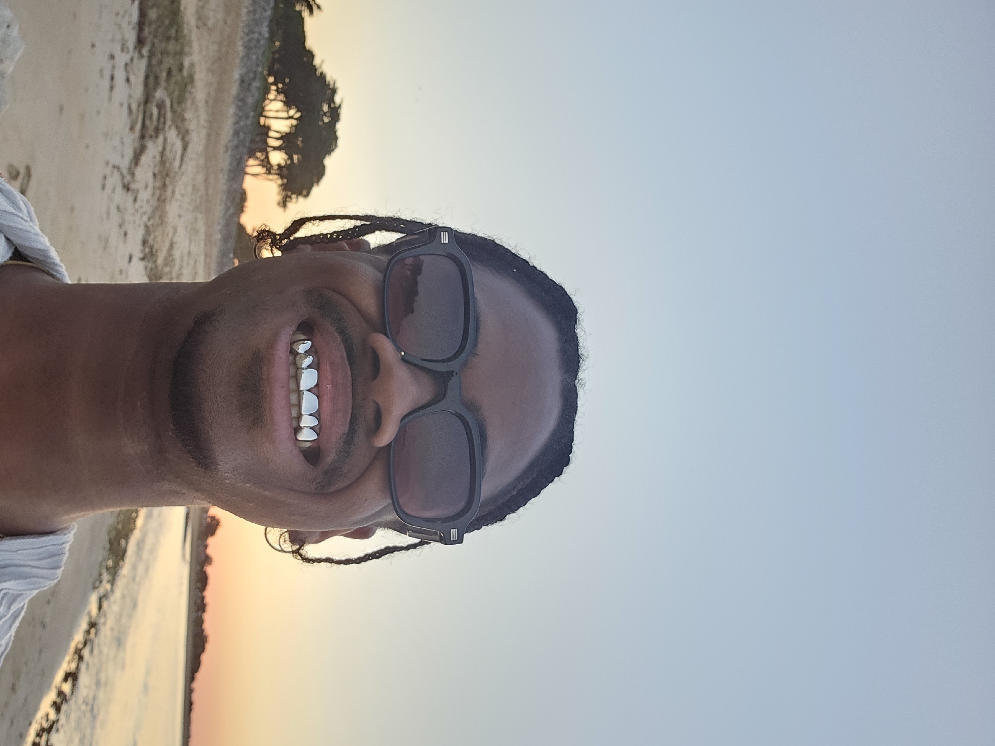 The image shows a man with dark skin, wearing black rectangular sunglasses and smiling to reveal silver teeth. He has braided hair and is wearing a white shirt. The background suggests a beach scene with sand, water, and trees silhouetted against a sunset sky. Given the prompt specifies the location as La Brée-les-Bains, France, the image can be described as a self-portrait taken by an individual on a beach in La Brée-les-Bains during sunset.
