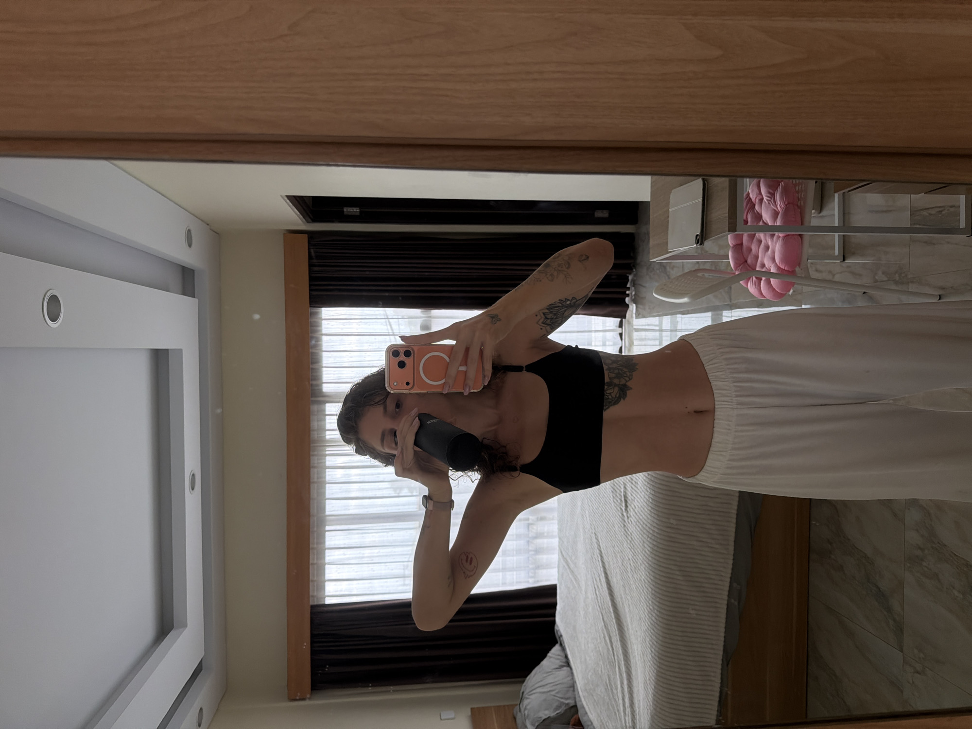 A woman is standing in a room, taking a selfie in a mirror. She is wearing a black sports bra and white wide-leg pants. She is holding a black water bottle to her mouth and taking a drink. Her phone, which has an orange case, is in her other hand, angled to capture the mirror's reflection. The room appears to be a bedroom, with a bed visible in the background. The walls are a light neutral color, and there are dark brown curtains covering a window. The floor is tiled. A wooden wardrobe is on the right side of the image, and a desk with a chair is visible in the background. The ceiling has recessed lighting and decorative trim. This scene is consistent with a residential interior in North Kuta, Indonesia.