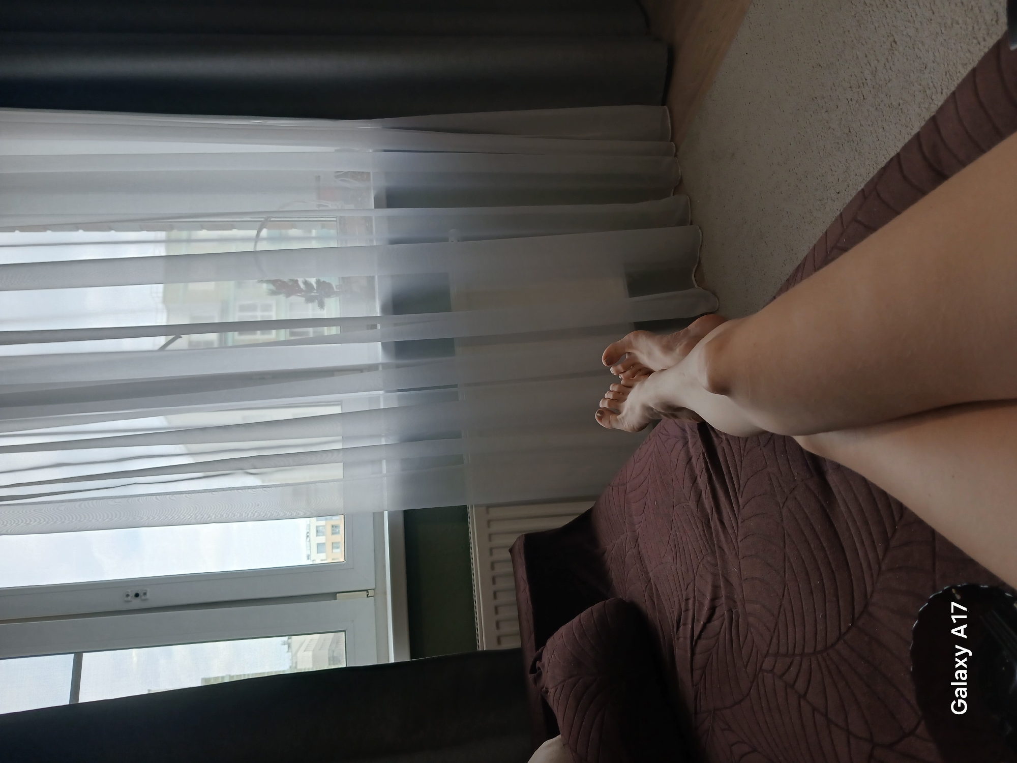 The image shows a person's legs resting on a brown patterned sofa. Behind the sofa, there's a window with white sheer curtains, through which buildings are visible, suggesting an urban environment. The phone's camera brand 'Galaxy A17' is visible in the bottom right corner of the image. Given the urban setting and the description, the location could be Yekaterinburg, Russia, considering that's part of the prompt.