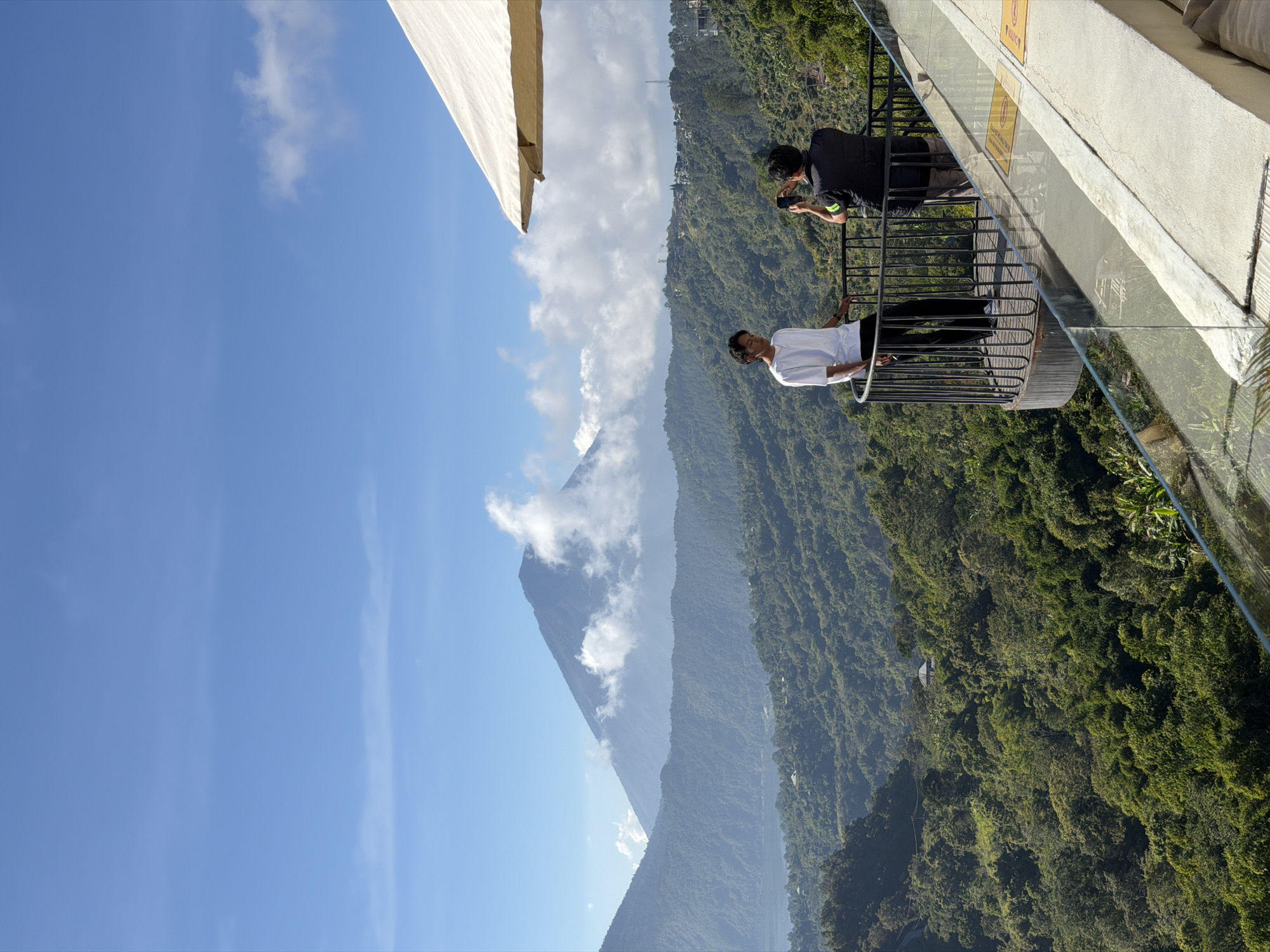 This is a vertical, medium shot photograph taken from a balcony in Kintamani, Indonesia, showcasing a vast, mountainous landscape under a bright blue sky with scattered white clouds. In the foreground, two individuals are standing on a curved balcony. One person, wearing a white t-shirt and black pants, is looking towards the left of the frame. The second person, dressed in a black vest over a dark shirt and grey pants, is facing away from the camera and holding up a phone to take a picture of the person in white. A large, light-colored umbrella is partially visible at the top right of the frame.
The middle ground is dominated by lush, green rolling hills and mountains covered in dense vegetation. A body of water, likely a lake, is visible in the distance on the left, nestled between the hills. Further in the background, a large, imposing volcano rises, its peak partially obscured by white clouds. The sky is a clear, vibrant blue with wispy cirrus clouds and some more substantial cumulus formations drifting around the volcano. The overall scene depicts a serene and scenic vista from a vantage point overlooking the natural beauty of Kintamani.