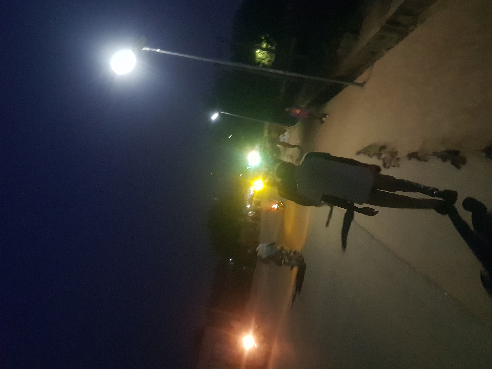 The image depicts a nighttime scene in what appears to be a residential area of Dabawa, Nigeria. Several streetlights illuminate the area. A group of people, including one individual in a light-colored shirt, are walking on a paved path. The ground is uneven with some debris. The background includes trees and a building.