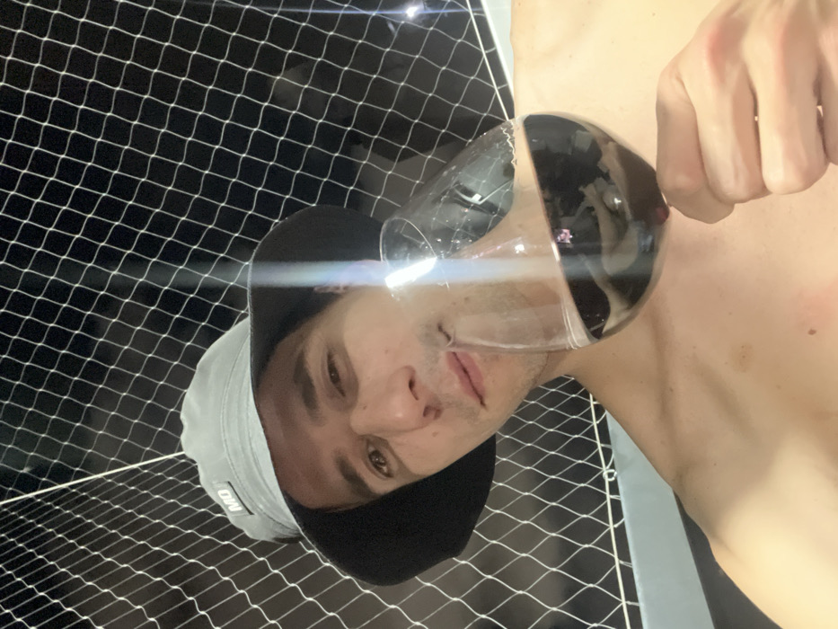 An adult male is depicted in a close-up, selfie-style shot, situated on what appears to be a residential balcony or terrace in an urban area of Buenos Aires, Argentina, at night. He is shirtless, with a light gray bucket hat featuring a black brim and a small "MO" logo patch on the front. He looks directly at the camera with a casual expression. His right hand holds a stemmed wine glass containing dark red liquid, positioned near his mouth as if he is about to drink or has just taken a sip. The background is dark, and a white, square-meshed safety net is prominently visible behind and around him, indicative of an elevated outdoor residential space.