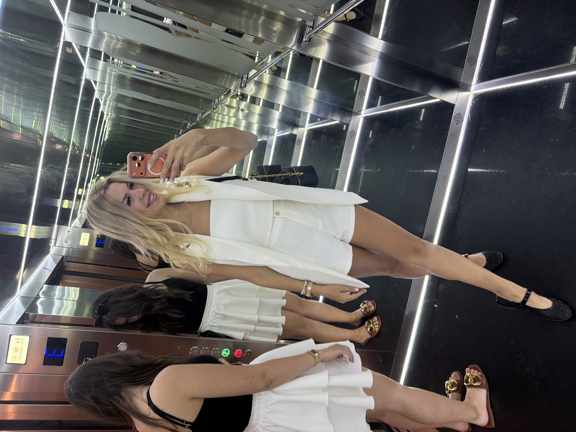 Three young women are inside a modern elevator with mirrored walls and a reflective black floor illuminated by white LED strips. The woman in the foreground, with blonde hair and wearing a white vest and shorts, is taking a selfie with her orange phone. To her left, a woman with dark hair wears a black top and a white tiered skirt, and to her left, another woman with dark hair is partially visible, also wearing a black top and a white tiered skirt. The elevator doors are visible in the background, with the letter "E" illuminated on a control panel. The scene appears to be in Polianytsia, Ukraine, likely within a resort or hotel given the modern and luxurious interior.