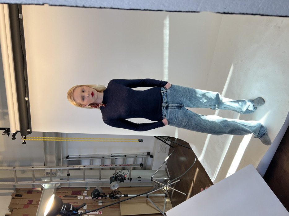 The scene depicts a photography studio located in Paris, France. A fair-skinned female model with blonde hair, red lipstick, and prominent red, multi-part earrings on her left ear stands centrally, facing the viewer. She is dressed in a dark blue, long-sleeved, high-neck top, acid-washed blue jeans with her hands in the pockets, and grey flat shoes. She maintains a neutral expression.
The background consists primarily of a white seamless paper backdrop that extends from the top of the frame to the floor, where it is also illuminated by natural light casting distinct rectangular shadows. To the left of the model, various studio equipment is visible: two strobe lights on stands, one larger with a silver reflector and diffuser and a smaller one below it. Further left, a tall metal ladder is positioned against a wall with stacked cardboard boxes containing labels such as "colorama" and "BD." A yellow chain hangs from the backdrop support system above. The floor to the immediate left of the backdrop is dark wood, and a white wall vent is visible. A white reflector card or bounce board is partially seen in the bottom left corner, held at an angle. The overall environment suggests an active commercial photoshoot setting.