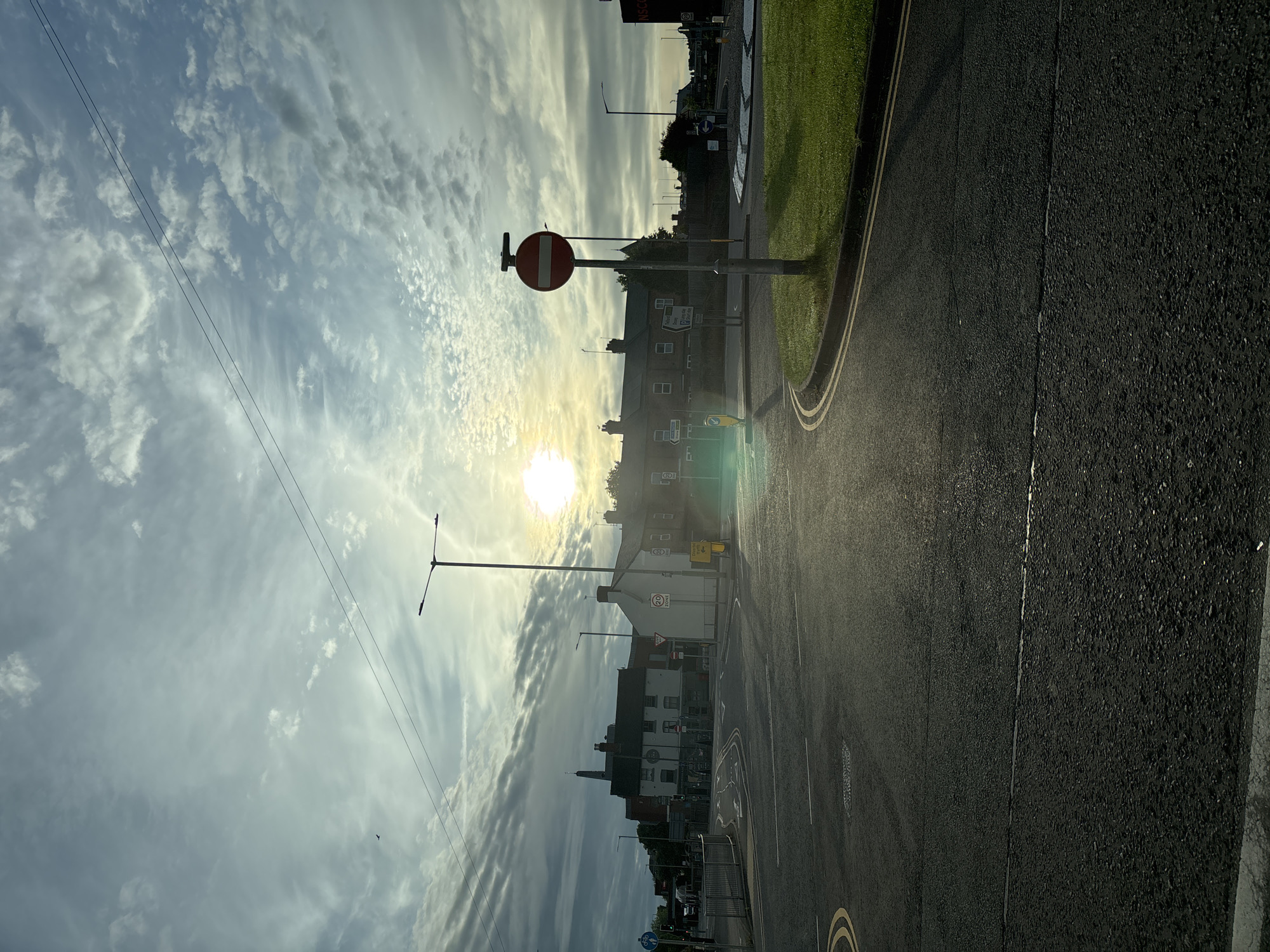 The image depicts a street scene in Stafford, United Kingdom, during what appears to be either sunrise or sunset, given the low angle of the sun and the bright glare. The perspective is from a road intersection, with asphalt paving and yellow double lines marking lanes. In the foreground, the road curves to the right, leading into a roundabout with a grass verge and a no-entry traffic sign prominently displayed on a pole.
Behind the roundabout, several buildings are visible, including a row of brick houses and a taller structure that might be a church spire or an older commercial building. Streetlights are present, and power lines stretch across the sky. The sky itself is a mix of blue and white clouds, with the sun creating a lens flare effect in the upper right quadrant.
Several traffic signs are discernible: a "20 ZONE" sign, a "GIVE WAY" sign, and a "NO ENTRY" sign with a red circle and a horizontal white bar. A yellow pedestrian crossing signal is also visible. In the distance, more buildings and streetlights are scattered across the landscape. A lone bird is captured in flight in the upper left portion of the sky. The overall impression is of a quiet, early morning or late evening in a suburban or town setting.