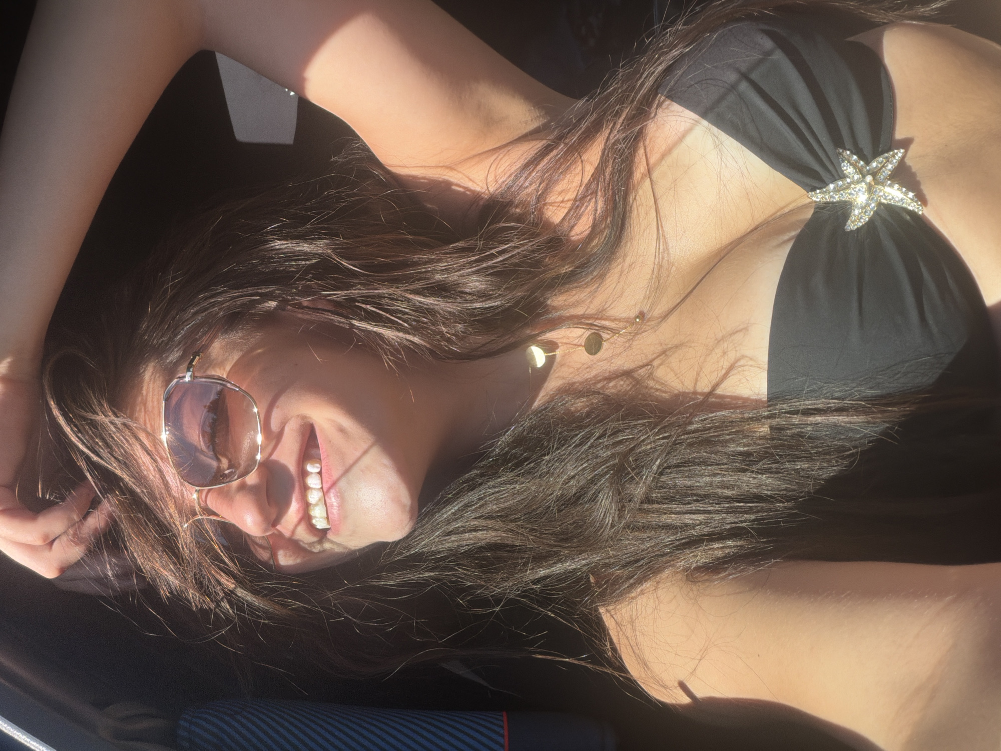 The image shows a woman wearing a black strapless bikini top adorned with a starfish-shaped embellishment. She has long, dark hair and is wearing sunglasses. The lighting suggests she is outdoors, possibly in a vehicle with the sun shining through a window, casting shadows on her face and body. The environment appears to be a bright, possibly coastal area, given the context of a bikini. The location in Burgas, Bulgaria, is not explicitly visible in the image itself, but it is stated as a known context.