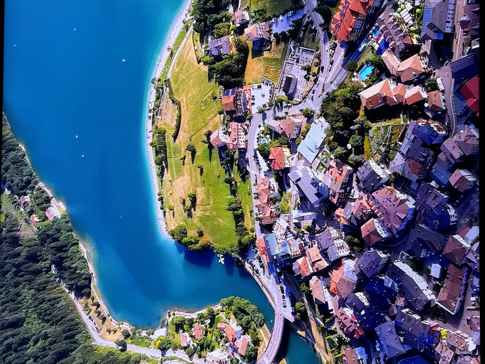 The image is an aerial view of a town situated alongside a bright blue body of water, likely a lake or a large river. The water has a distinct turquoise hue and is bordered by a sandy or light-colored shoreline. On the land, a dense green forest covers a hillside that slopes down towards the water.
Adjacent to the water and the forested area, a settlement is visible, characterized by a dense cluster of buildings with terracotta-colored roofs. These buildings are of varying sizes and architectural styles, many featuring multiple stories. Some structures appear to be residential homes, while others might be businesses or hotels, judging by their arrangement and proximity to the water and a road.
A paved road winds through the scene, connecting different parts of the town and leading towards the water. Several vehicles are parked along the roadside and within the town. In the grassy areas between the buildings and the water, there are signs of recreational activity, with what appear to be sun loungers and umbrellas set up on the beach. Small white specks on the water suggest the presence of boats or watercraft. The overall impression is a picturesque European alpine town with a scenic waterfront. This description does not relate to Chirchik, Uzbekistan.
