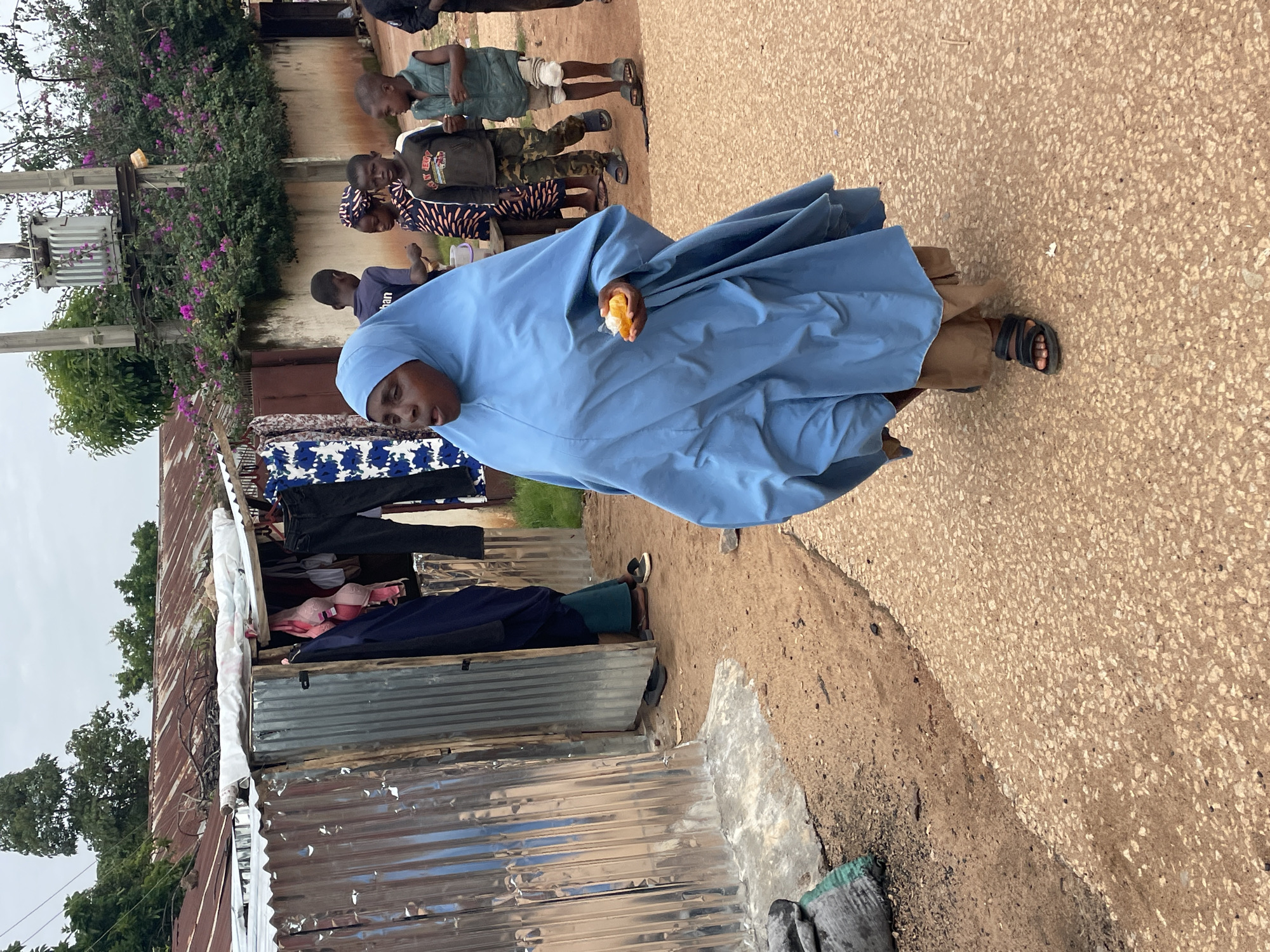 In a bustling street scene in Jos, Nigeria, a woman in a blue hijab walks briskly, holding a piece of orange fruit. She is surrounded by a group of children and young people. Behind them, corrugated metal shacks line the dusty road, some displaying clothing for sale. A transformer hums on a pole, adorned with vibrant purple flowers. The atmosphere is lively, with a mix of commerce and daily life unfolding under a cloudy sky.