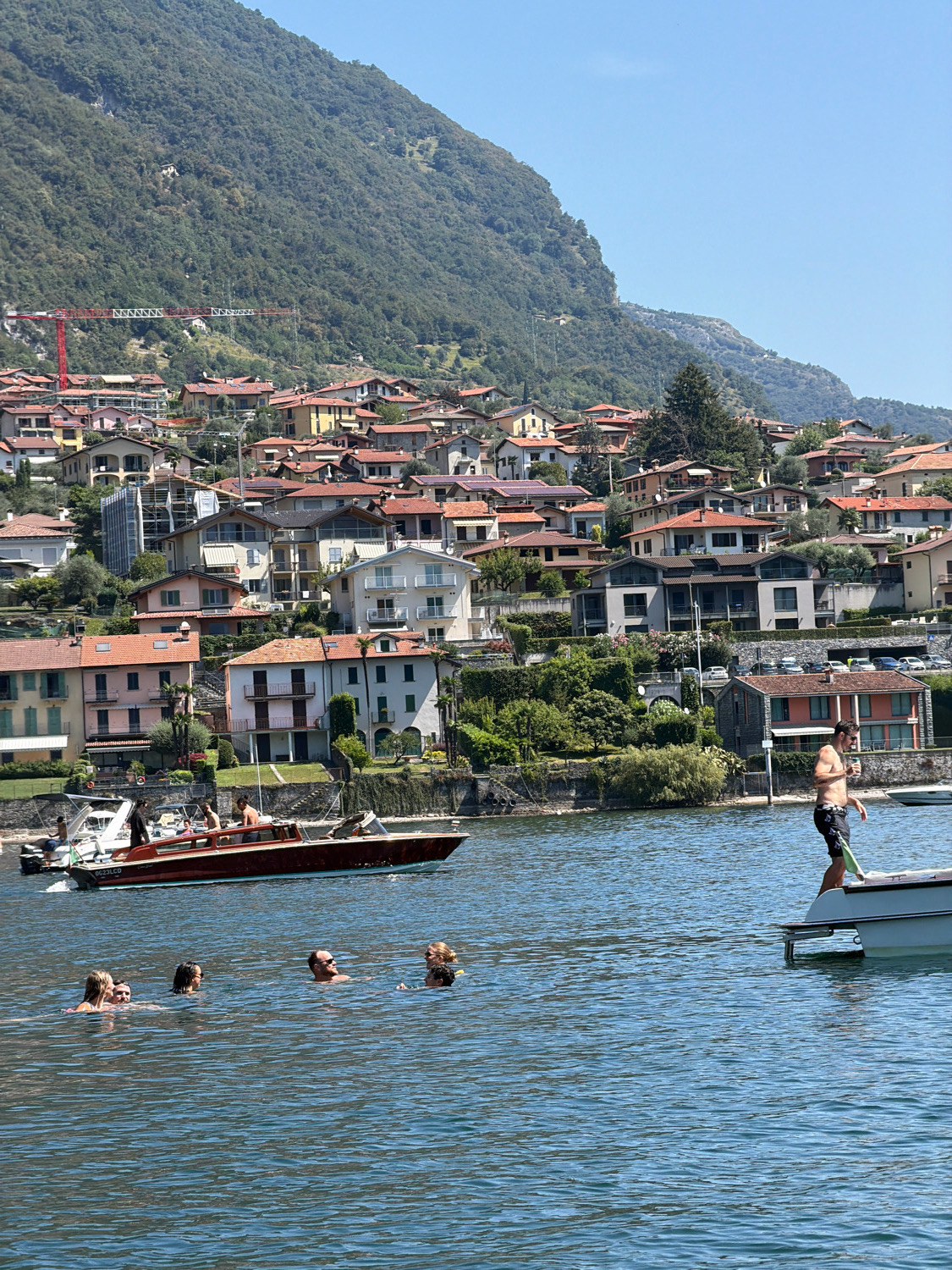 The image depicts a sunny day on Lake Como in Tremezzina, Italy. In the foreground, a group of five people are swimming in the clear blue water. To the right, a man stands on the edge of a white boat, holding a green object. In the middle ground, a long, wooden boat with several occupants is moving across the lake. The background features a hillside covered with lush green trees, densely packed with residential buildings and homes with red-tiled roofs, characteristic of Italian lakeside towns. A red construction crane is visible on the hillside, suggesting ongoing development. The sky is a clear, bright blue.