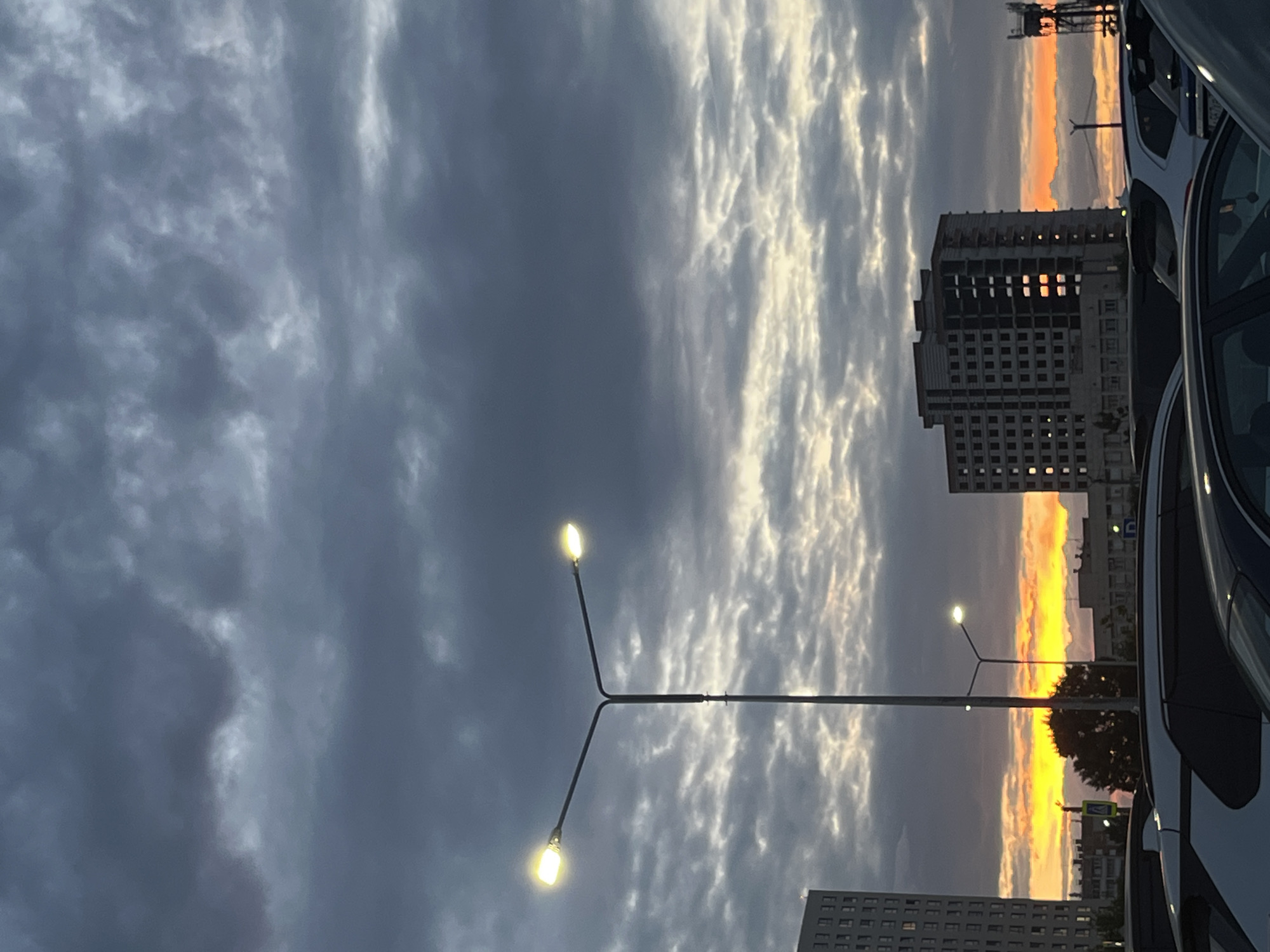 This media file captures a street scene in Chelyabinsk, Russia, during what appears to be dusk. The composition is dominated by a dramatic sky filled with clouds, some dark and heavy, others illuminated by the setting sun, creating a striking contrast. The sky displays hues of grey, white, and vibrant orange and yellow near the horizon, suggesting either a sunset or sunrise.
Prominent in the foreground are two streetlights, their bulbs casting a warm, yellow glow against the darkening sky. They are mounted on a metal pole that extends horizontally across the frame.
In the mid-ground, a multi-story building with a facade of numerous windows stands. The windows reflect the bright sky, adding to the visual interest. To the right of this building, a vehicle is partially visible, its dark frame and windows suggesting it's parked or moving slowly. Further in the background, another building and a tree are silhouetted against the colorful sky. The presence of streetlights and buildings indicates an urban setting.
The overall mood is serene yet dramatic, with the interplay of light and shadow, and the contrasting elements of urban infrastructure against a natural sky phenomenon. The time of day is clearly indicated by the sky's coloration and the illuminated streetlights, marking the transition from day to night. No visible text is present in the media file.
