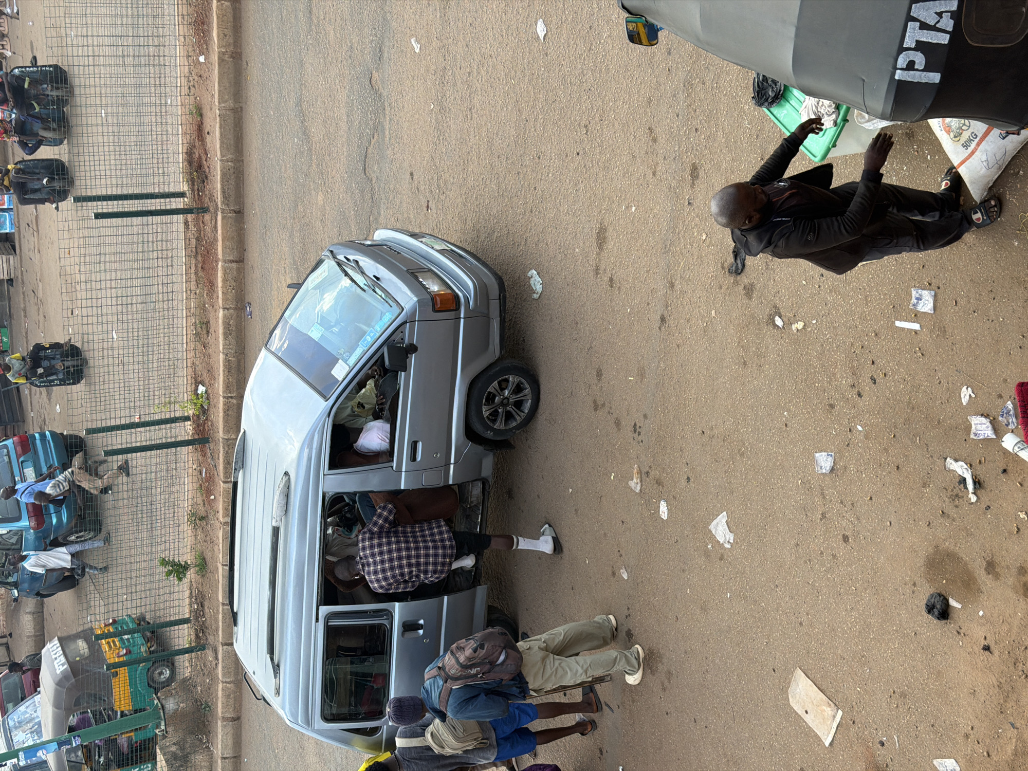 The image depicts a street scene in Bukuru, Nigeria. In the foreground, a man in a black tracksuit and sandals stands near a green plastic container and a large bag labeled "50KG". To his right, a part of a vehicle with the letters "PTA" visible on its side is present. Behind this, a silver van is parked with its door open. Several people are visible around the van. One man wearing a plaid shirt is exiting the van, while another man in a blue shirt and backpack stands to the left of the van. A third person, wearing a blue shirt and a grey beanie, also has a backpack. Further in the background, other vehicles, including a blue minivan and a tricycle, are parked on the street, separated by a wire fence. The ground is paved with asphalt and shows signs of wear and litter.