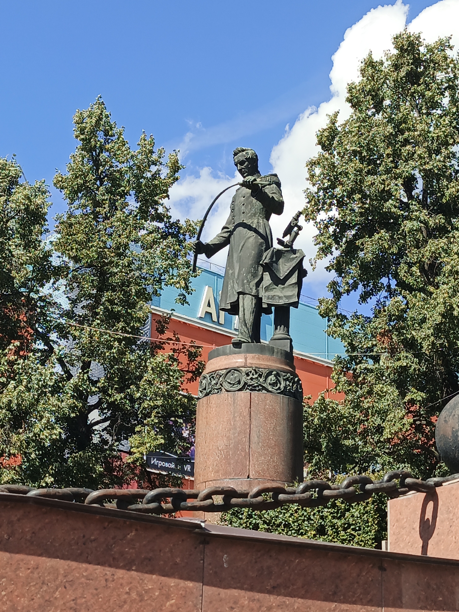 The image shows a bronze statue of a man in a military uniform holding a curved sword. The statue is on a pedestal made of granite and adorned with intricate carvings. Behind the statue, a vibrant blue and orange building is visible, partially obscured by lush green trees. In the foreground, a heavy metal chain stretches across the frame, suggesting a boundary or fence. The sky above is a clear blue with scattered white clouds. The statue is located in Zlatoust, Russia.