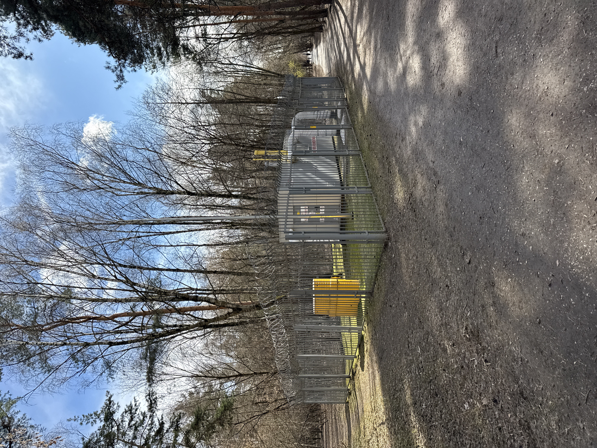 The image shows a fenced-in utility facility located alongside a dirt path in a wooded area. The facility appears to be metallic gray and yellow in color, and surrounded by a gray chain link fence, topped with barbed wire. Trees with bare branches surround the fence. The path is dirt and slightly inclined. Sunlight filters through the trees, creating shadows on the ground. The sky is visible above the trees, with patches of blue and some clouds. It is difficult to pinpoint the exact location within Moscow, Russia, without further information or context.