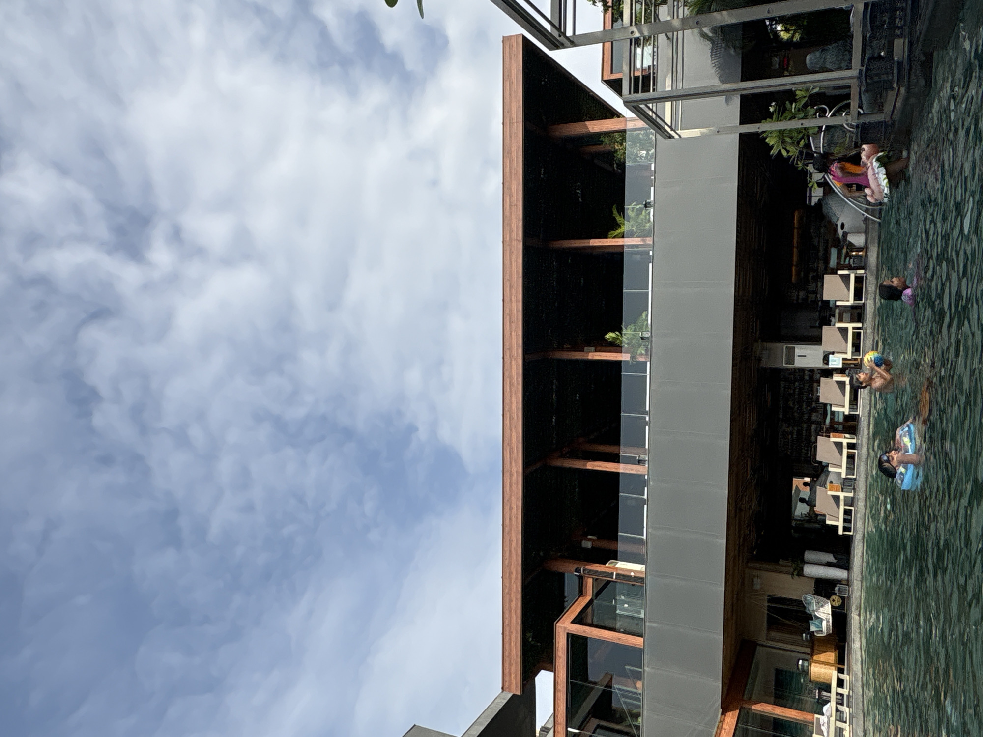 The image captures a scene at a resort or hotel in Vagator, India, featuring a swimming pool in the foreground and a multi-story building with a contemporary design in the background. The sky above is filled with scattered white clouds against a light blue expanse.
In the pool, three children are visible. One child is wearing a blue float and appears to be looking towards the camera. Another child, also in the pool, is partially visible from the side, holding a yellow object. A third child, dressed in pink and orange, is partially submerged, holding onto a float with a patterned skirt.
The building in the background exhibits a modern architectural style with a prominent dark-colored roof supported by wooden beams. The upper level features a balcony with glass railings, overlooking the pool area. Below the balcony, the lower level of the building appears to be an open-air space with furniture, including lounge chairs and tables, suggesting a dining or relaxation area. The building's exterior incorporates large glass windows and dark cladding. Tropical foliage is present around the structure, indicating a warm climate. The overall impression is one of a leisurely setting, likely a holiday destination.