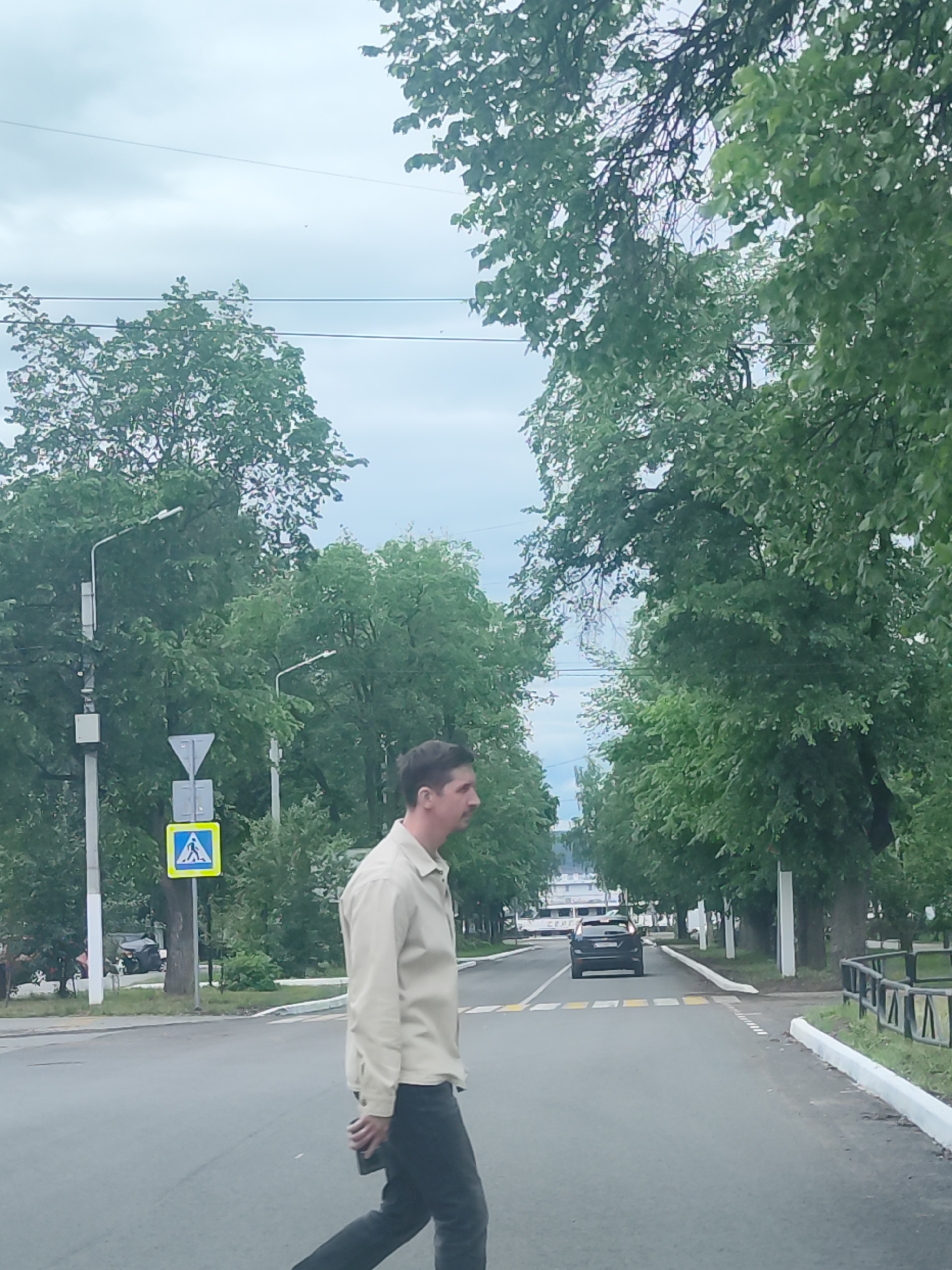 A man with a mustache walks across a crosswalk on a street in the city of Chaikovsky, Russia. He wears a tan shirt and dark pants, and holds a dark object in his hand. A black car approaches the crosswalk. A pedestrian crossing sign is visible on the left side of the road. Green trees line the sides of the street. The sky is overcast.