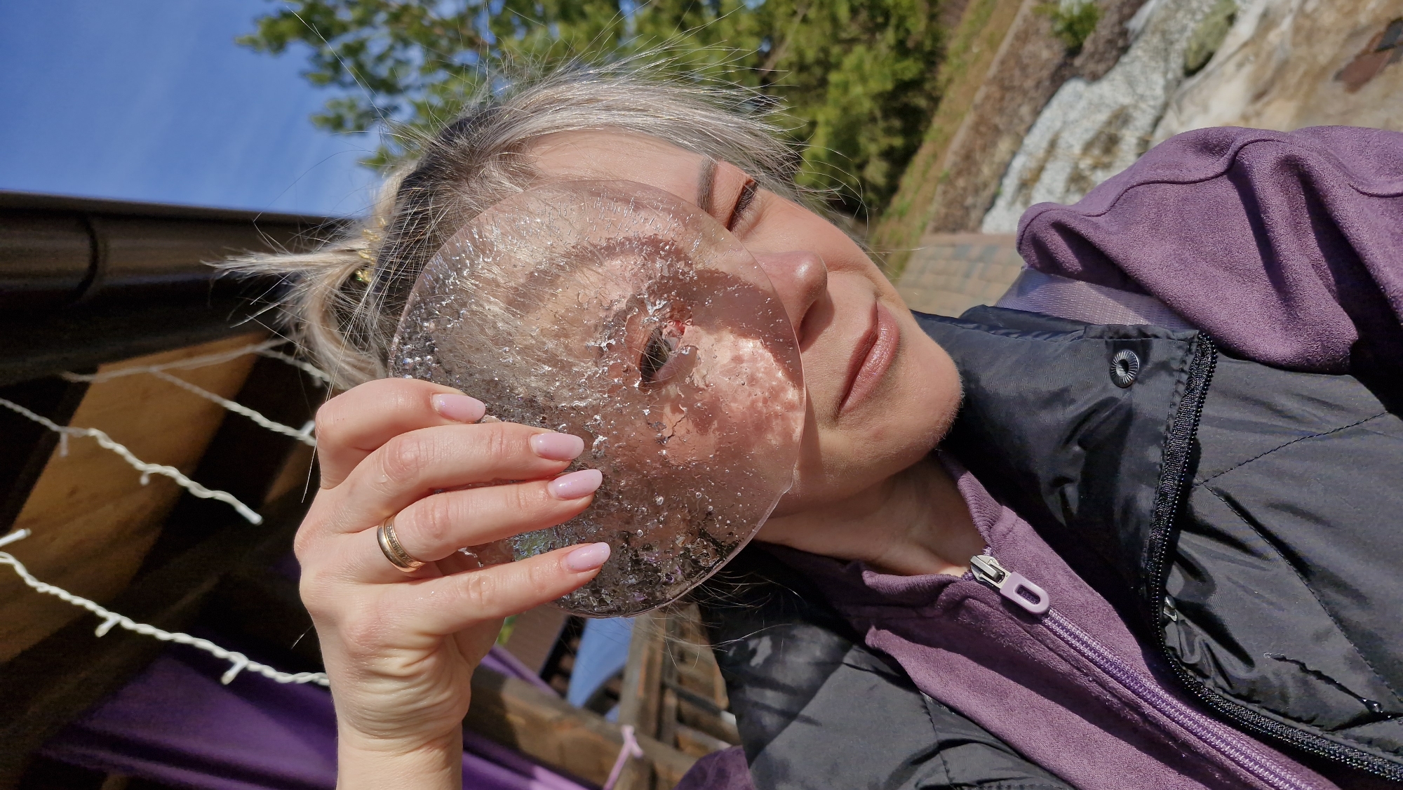 A woman with blonde hair and fair skin is holding a clear, circular object, possibly ice, in front of her face. She is wearing a gold ring on her left ring finger and has pink nail polish. She is dressed in a purple hoodie and a black jacket. The background shows a blurred outdoor scene with greenery, a blue sky, and possibly a wooden structure with white lights. The location appears to be outdoors, and although the specific location within Otchor, Russia, cannot be determined from the image, the presence of greenery and a clear sky suggest a natural or park-like environment.