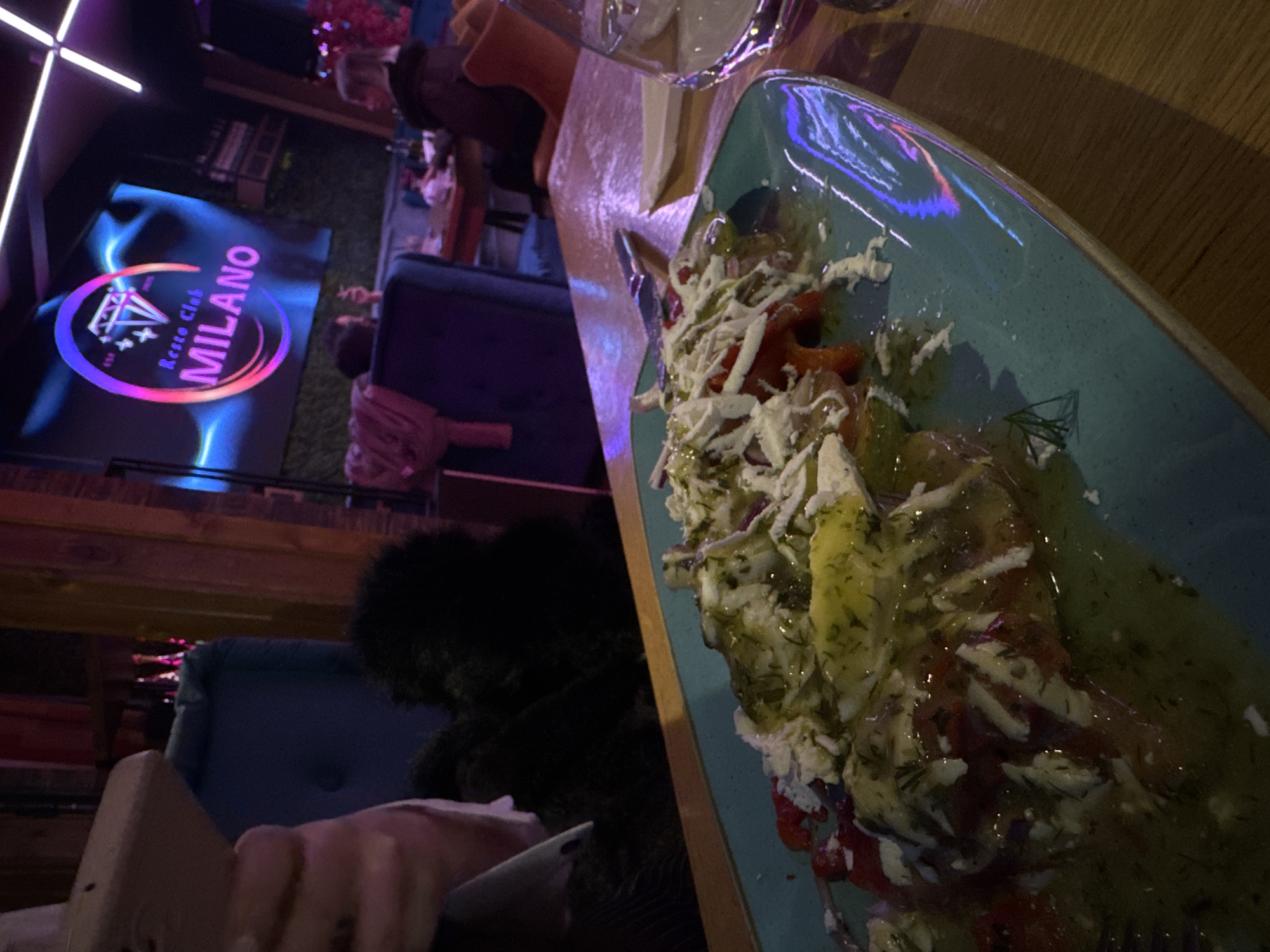 A low-angle shot inside a restaurant or club, possibly Milano Resto Club in Sofia, Bulgaria, based on the branded logo on a screen in the background. The focus is on a plate of food, likely a salad, on a wooden table. The salad consists of greens, shredded white cheese, red peppers, and a dressing or sauce. The plate is teal-colored with a glazed finish. A glass, partially visible, is on the table. A person is seen holding a mobile phone or similar device in their left hand. The background includes blurred figures, the restaurant’s interior, lighting fixtures, and the screen displaying the Milano Resto Club logo. The environment appears dimly lit, and the overall composition is angled, suggesting the image was taken spontaneously.