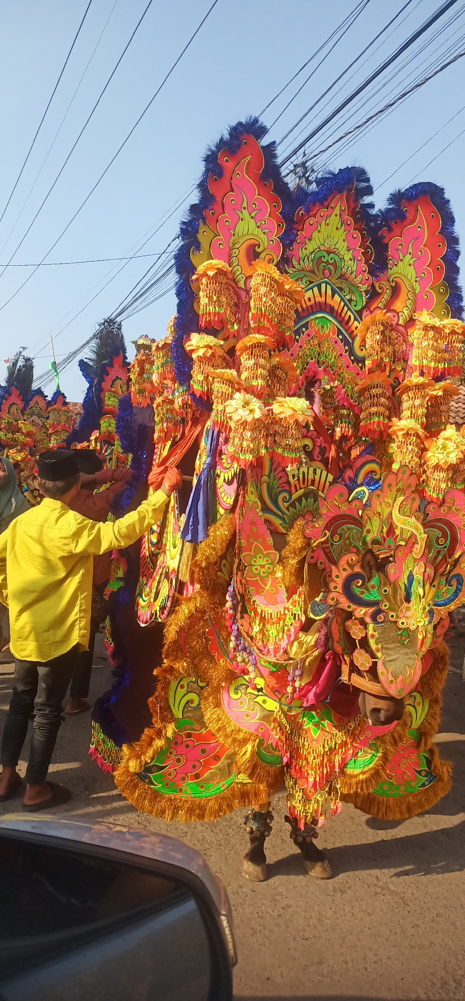 The image depicts a vibrant cultural parade in Kalisalam, Indonesia. The central focus is a performer in an elaborate, colorful costume resembling a mythical creature, possibly a dragon or a phoenix. The costume is adorned with numerous shimmering ornaments, intricate patterns in shades of pink, orange, green, and blue, and a prominent display of tassels and beadwork. The performer's head is concealed within the costume, but their lower body is visible, featuring hoof-like structures and ankle adornments. A man in a bright yellow shirt and black pants, wearing a black cap, stands to the left of the performer, interacting with the costume, possibly adjusting it or participating in the parade. Other individuals are partially visible in the background. The environment appears to be an outdoor street scene under a clear blue sky, with overhead power lines suggesting an urban or semi-urban setting. A car's side mirror is visible in the foreground, reflecting the scene.