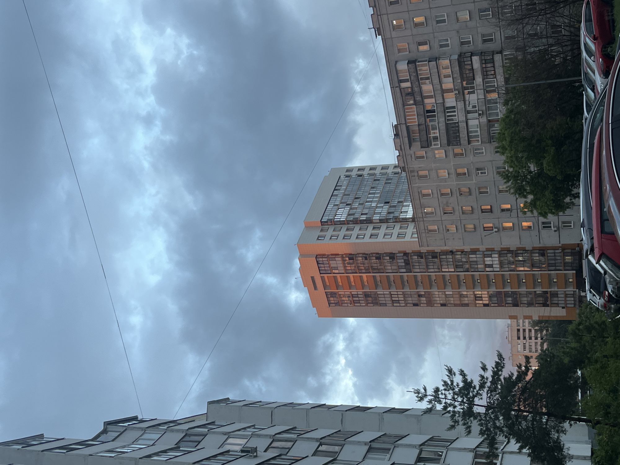 The image captures an urban residential scene in Kazan, Russia, under a cloudy, overcast sky, likely during dusk or early evening as indicated by illuminated windows in some buildings. Several multi-story apartment buildings are the primary subjects.
To the right, an older, gray residential block with numerous small windows and enclosed balconies lines the frame, with lights visible in some units. Alongside this building, a row of parked cars, including a prominent red one, suggests a street or parking area. In the center, a taller, more modern high-rise building stands out with a facade featuring a mix of reflective windows and white and orange-hued panels. This building also displays internal lights. Another residential structure, with a distinct roofline, is partially visible in the lower left.
Various trees are interspersed throughout the scene, providing greenery, with a notable conifer tree in the lower-middle section. Thin power lines stretch across the gray sky, adding a utilitarian element to the composition. The overall mood is quiet and everyday, typical of a city at the end of the day. No people or specific activities are depicted, and no visible text is discernible.
