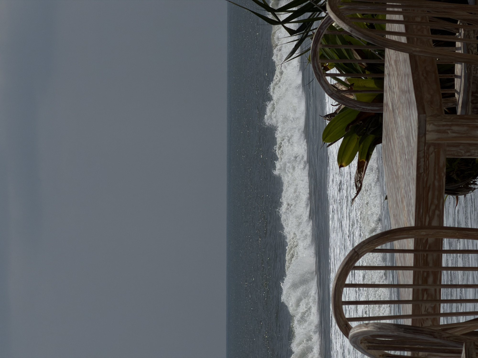 The image depicts an unoccupied beachfront setting in Belalang, Indonesia. In the foreground, a light-toned wooden table occupies the lower central and right portions of the frame. Two matching light-toned wooden spindle-back chairs are partially visible, one prominently on the left and another to the right, behind the table. Green tropical foliage, featuring broad leaves and some dry sections, is positioned around and behind the table and chairs, primarily on the right side. The midground features the ocean, presenting a dark blue-grey surface with visible texture from water movement. A significant white wave is actively breaking in the central horizontal plane, generating substantial foam and whitewater. The background is dominated by a uniformly overcast grey sky, which meets the ocean at a distinct horizon line. No individuals are present in the scene.