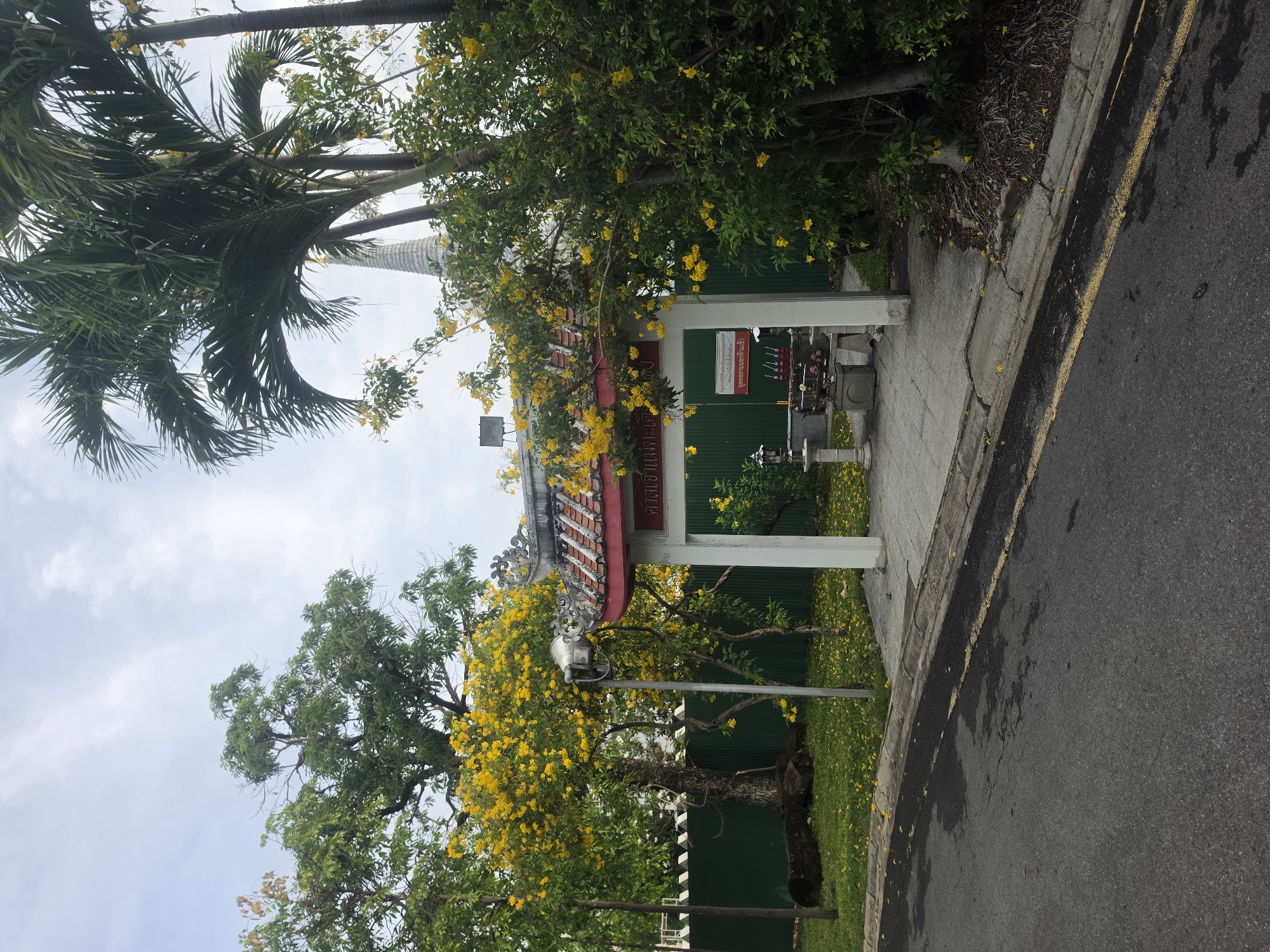 The image shows a street scene in Bangkok, Thailand, featuring a small, ornate, shrine-like structure with a red-tiled roof supported by white columns. The shrine is located on a paved area adjacent to a road with a yellow line marking the curb. Lush green foliage, including trees with vibrant yellow flowers, surrounds the shrine. The background includes a green fence and glimpses of other buildings and trees. The sky is visible in the upper portion of the frame, with scattered clouds.