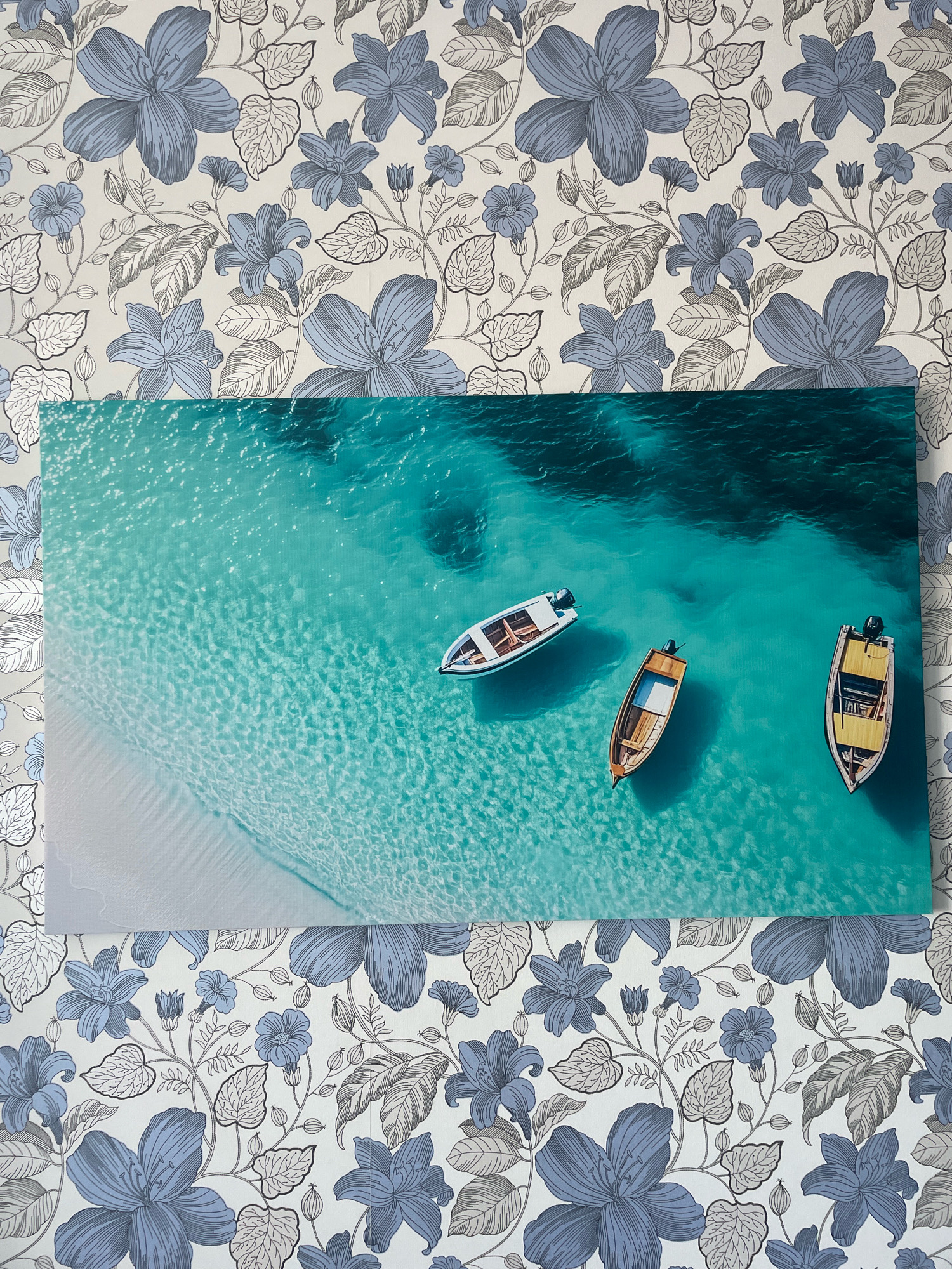 The image shows a framed artwork of three small boats floating in clear turquoise water near a sandy beach. The artwork is displayed against a floral wallpaper featuring blue and gray flowers and leaves. There are no people or animals visible in the image, and no specific identifiable location within Krasnodar, Russia, is depicted. The scene is static and appears to be an indoor setting.