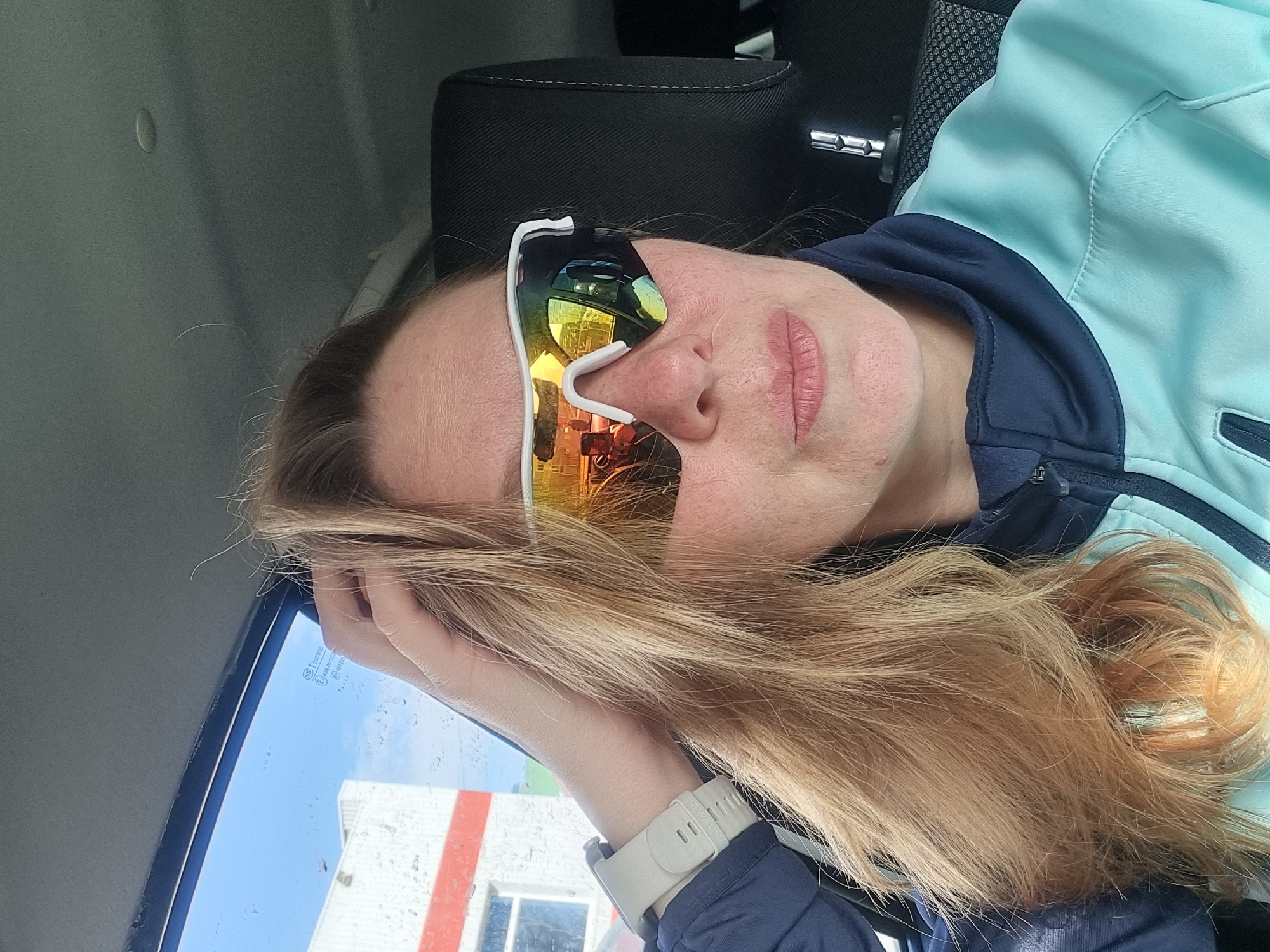 A female subject, appearing Caucasian and possibly in her 30s or 40s, is depicted inside a vehicle. She has long, light brown hair, some strands of which are unruly. She wears white-framed sports sunglasses with reflective, multi-hued lenses (predominantly orange, yellow, and blue). Her lips are natural pink. A light-colored smartwatch with an "EWADE" branded band is visible on her left wrist. She is dressed in a two-tone athletic jacket, light blue (teal) on the main body and dark blue on the collar and shoulder sections. Her right hand is raised, fingers lightly touching her hair near her right temple.
The interior of the vehicle includes a grey headliner and a black headrest with visible white stitching behind her right shoulder. Through the vehicle's side window, which is speckled with water droplets, an exterior building is visible. This building is predominantly white with a prominent horizontal red stripe positioned high on its facade. A window opening is also visible on the building. The sky above the building is blue. Reflections within the sunglasses show elements of the vehicle's interior and a partial reflection of another individual or object with an orange hue. The subject is oriented looking slightly to her right, away from the immediate camera perspective.
The specific location within the city of Chaikovsky, Russia, cannot be determined from the generic architectural features (white building with a red stripe) visible through the car window. No distinct landmarks or street identifiers are present.