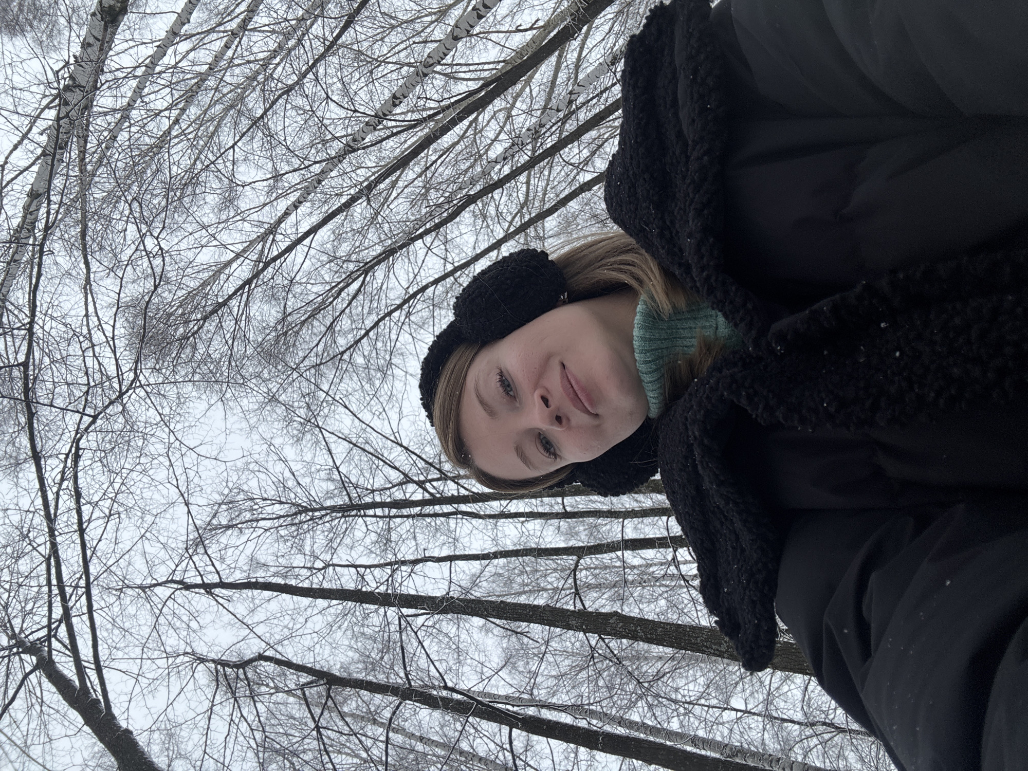 The image is a selfie taken from a low angle, looking up towards a woman in winter clothing with bare birch trees in the background. The woman is wearing black earmuffs, a green sweater, and a large black puffer jacket with a dark, textured scarf or shawl around her neck. Her face is visible, and she is smiling slightly. The background is filled with the stark, bare branches of birch trees against a pale sky, suggesting a winter setting. The location is possibly in a park or wooded area within the city of Chaikovsky, Russia, given that this query was from that location. The primary action is the woman taking a selfie. There is no visible interaction with other people or subjects. The environment is a cold, snowy wooded area in winter.