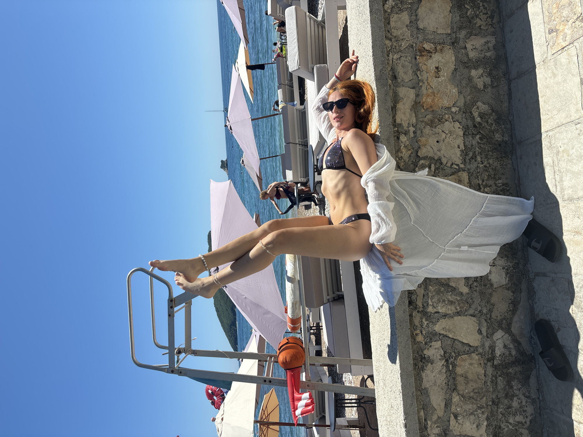 A young woman with auburn hair is shown reclining on a white lounge chair at a sunny beachside location in Budva, Montenegro. She wears a sparkly black bikini and a sheer, flowing white cover-up, with dark sunglasses on her face and pearl-like anklets on her bare feet. Her legs are elevated, resting on the metal frame of a lifeguard stand, giving a relaxed pose as she looks towards the camera.
The scene is set against a vibrant blue sky and the calm, clear turquoise waters of the sea. In the background, numerous white lounge chairs with grey cushions and light-colored beach umbrellas line the shore, with other people enjoying the sun and water. A distant landmass is visible across the water. Objects like a red and white striped buoy and an orange float are present near the waterline, along with a red parachute-like device possibly related to watersports. The foreground features a textured stone wall and ground, where a pair of black slide sandals are placed. The strong, direct sunlight and pronounced shadows indicate it is likely midday or early afternoon. The overall atmosphere is one of leisure and enjoyment at a vibrant coastal resort. There is no visible text in the image.