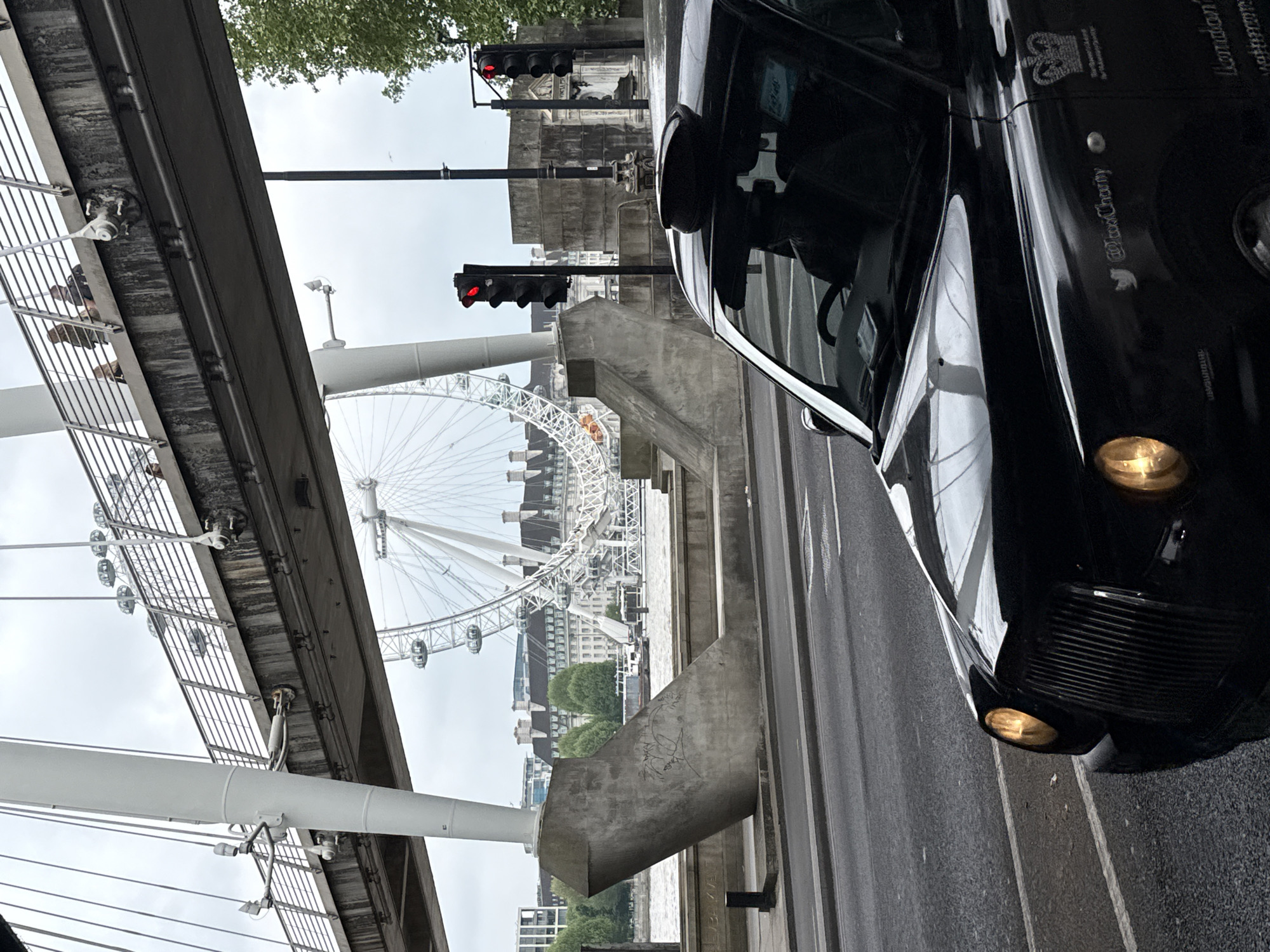 The image shows a view of the London Eye, a large Ferris wheel, seen through the infrastructure of a bridge. The bridge appears to be a pedestrian bridge with metal railings. The scene is taken from a street level, with the front of a black London taxi prominently featured on the right side of the frame. Traffic lights are visible, showing red. The sky is overcast, suggesting a cloudy day. The environment includes buildings in the background. The image is somewhat tilted.