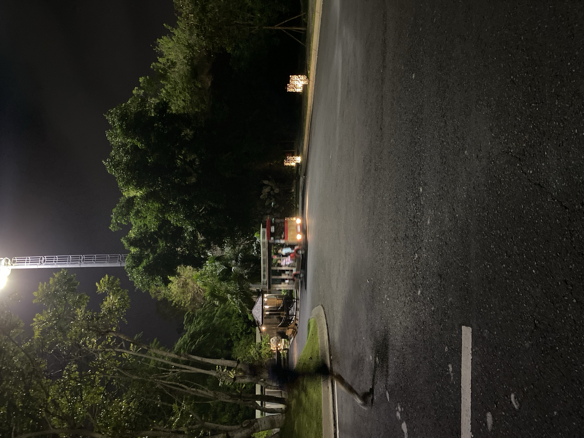 In Kuta Selatan, Indonesia, at night, an asphalt road curves gently to the right, bordered by a manicured grassy verge on the left and dense foliage and trees on the right. A tall, illuminated tower stands in the background, its light casting a glow on the surroundings. On the road, a small, open-air vehicle, resembling a tram or shuttle, with a red and yellow exterior, is in motion, carrying several passengers. To the left, a person is captured in motion, appearing as a blur, running or walking across the road. Scattered along the right side of the road are decorative lights with intricate cut-out patterns, illuminating the pathway. The scene suggests a resort or entertainment venue illuminated for evening activity.