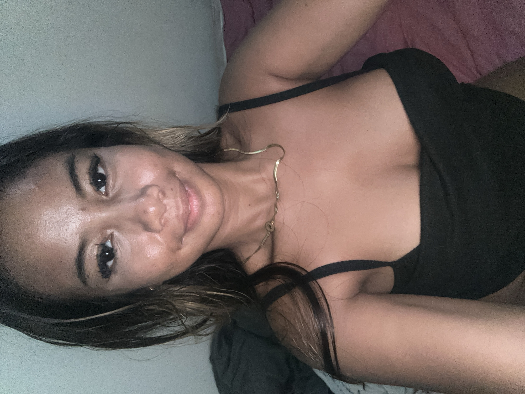 A woman is lying down, facing the camera. She is wearing a black top and a gold necklace. The background appears to be a bedroom setting with a bed and wall. The location of the photograph is not possible to confirm as being taken in Sukawati, Indonesia.
