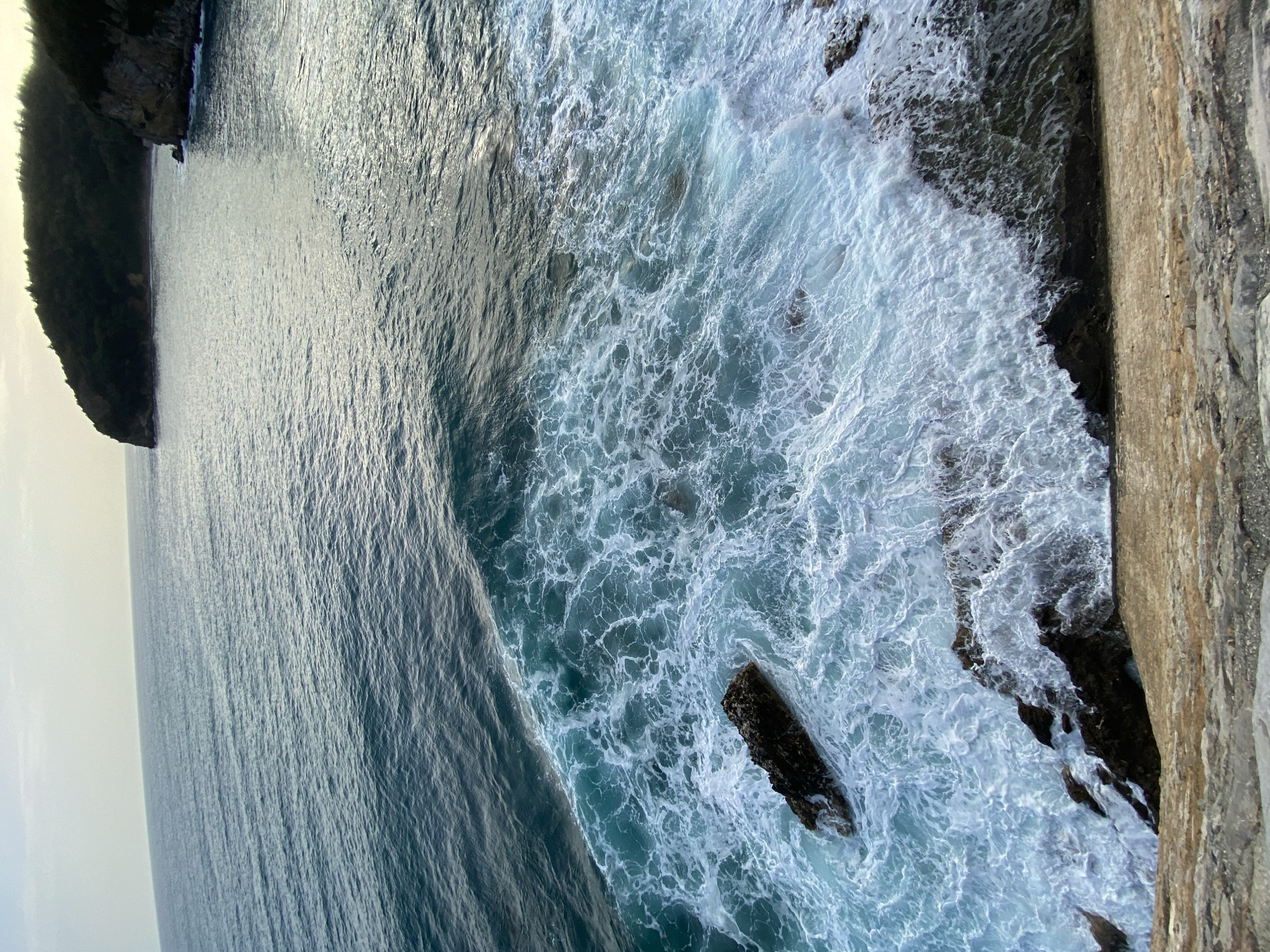 The image shows a view from a high vantage point looking down at the Adriatic Sea along the coast near Budva, Montenegro. To the left, a dark, forested headland juts out into the sea. The sea transitions from a calmer, dark blue to a more turbulent, light blue-white as it approaches the rocky shoreline on the right. White foamy waves are crashing against the rocks and part of a stone wall is visible on the extreme right edge of the image, indicating the viewpoint is likely from a structure like a cliffside path or building.