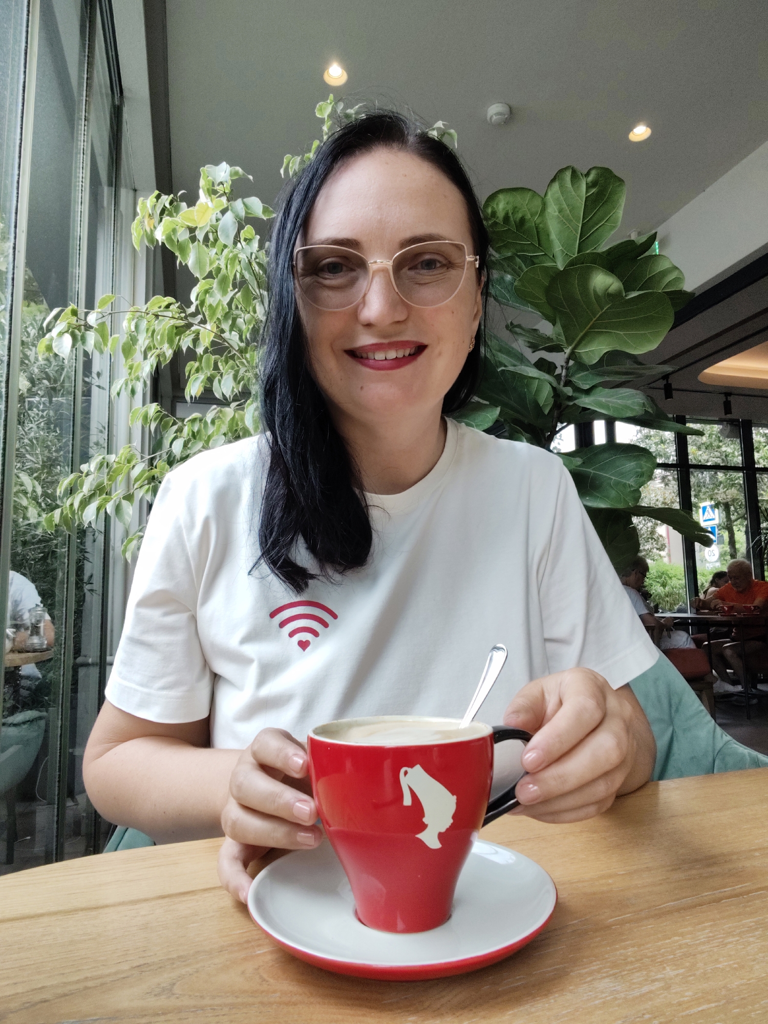 A woman with dark hair and glasses smiles at the camera, holding a red mug of coffee with a spoon. She is wearing a white t-shirt with a red Wi-Fi symbol and a heart printed on it. She is seated at a wooden table in what appears to be a cafe with large windows and plants visible in the background. The location is identified as Sochi, Russia. Other patrons are visible in the background, seated at tables. The overall scene depicts a casual moment in an urban cafe setting.