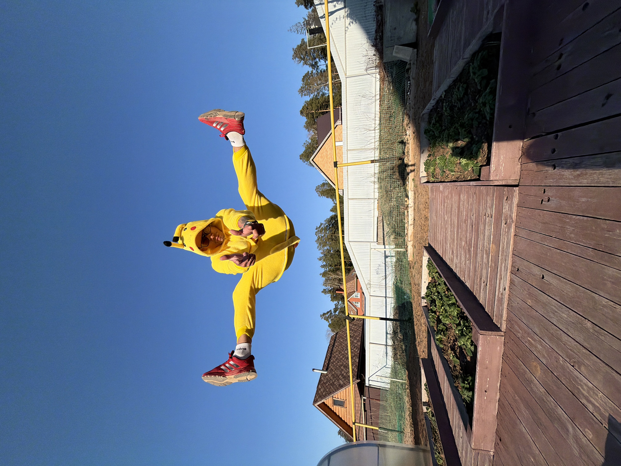 A person wearing a Pikachu costume and red Adidas shoes is performing a split in the air. The background shows a clear blue sky on one side, and a fenced residential area with wooden structures and greenery on the other. The wooden structure appears to be a deck with plant beds. While a precise location within Chaikovsky, Russia, is impossible to determine from the image alone, the style of buildings and fencing is consistent with residential areas in Russia.