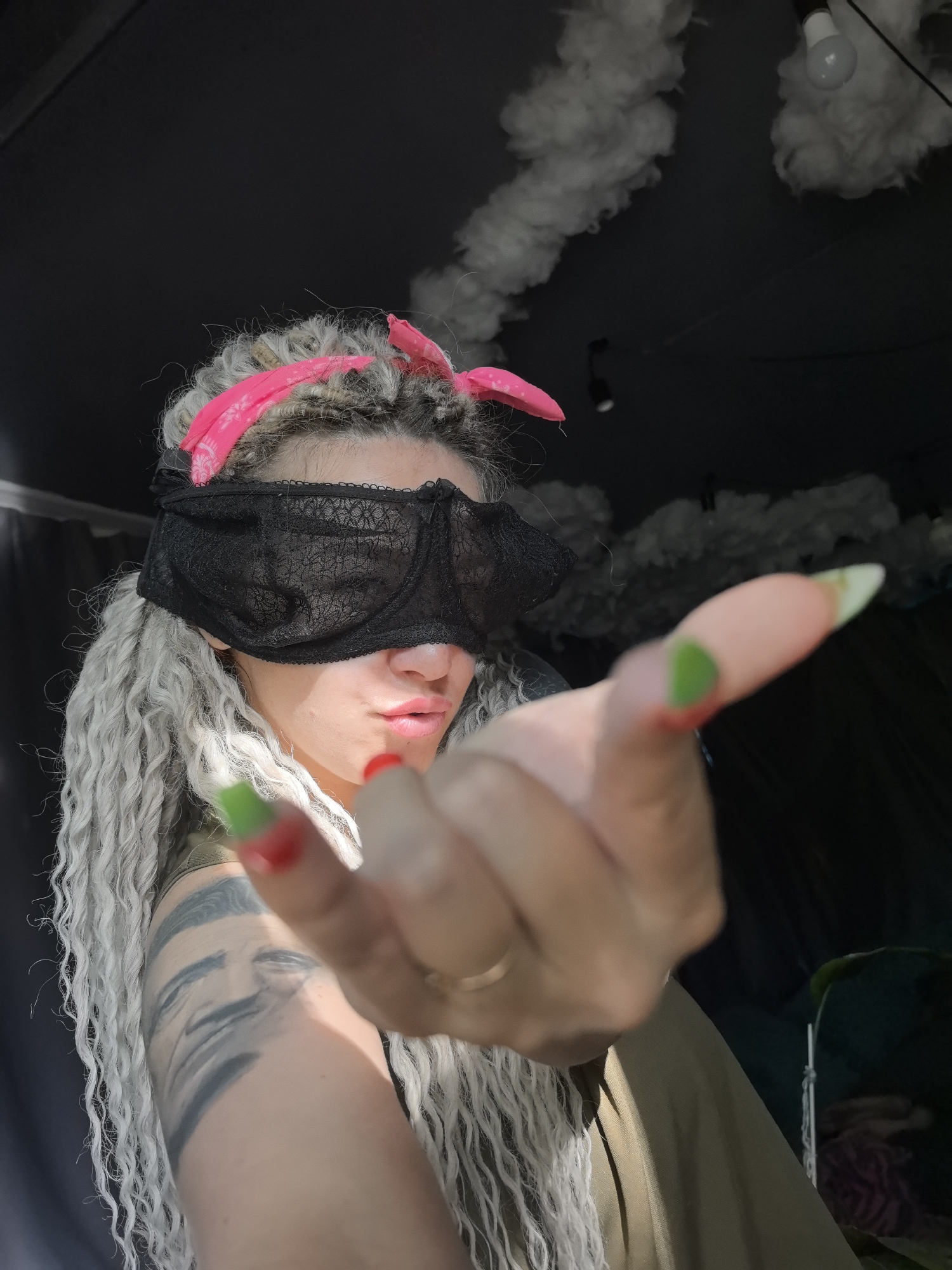 A woman is posing indoors with a black lace bra acting as a blindfold over her eyes. She has long, curly blonde hair with a pink bandana tied around her head. Her left arm is extended towards the camera. She has a tattoo of a face on her left arm. Her nails are painted green and red. The background includes dark curtains, artificial clouds, and a light bulb hanging from the ceiling. The location is likely within an indoor space in an unknown location, with no information to indicate the city of Noviy, Russia.