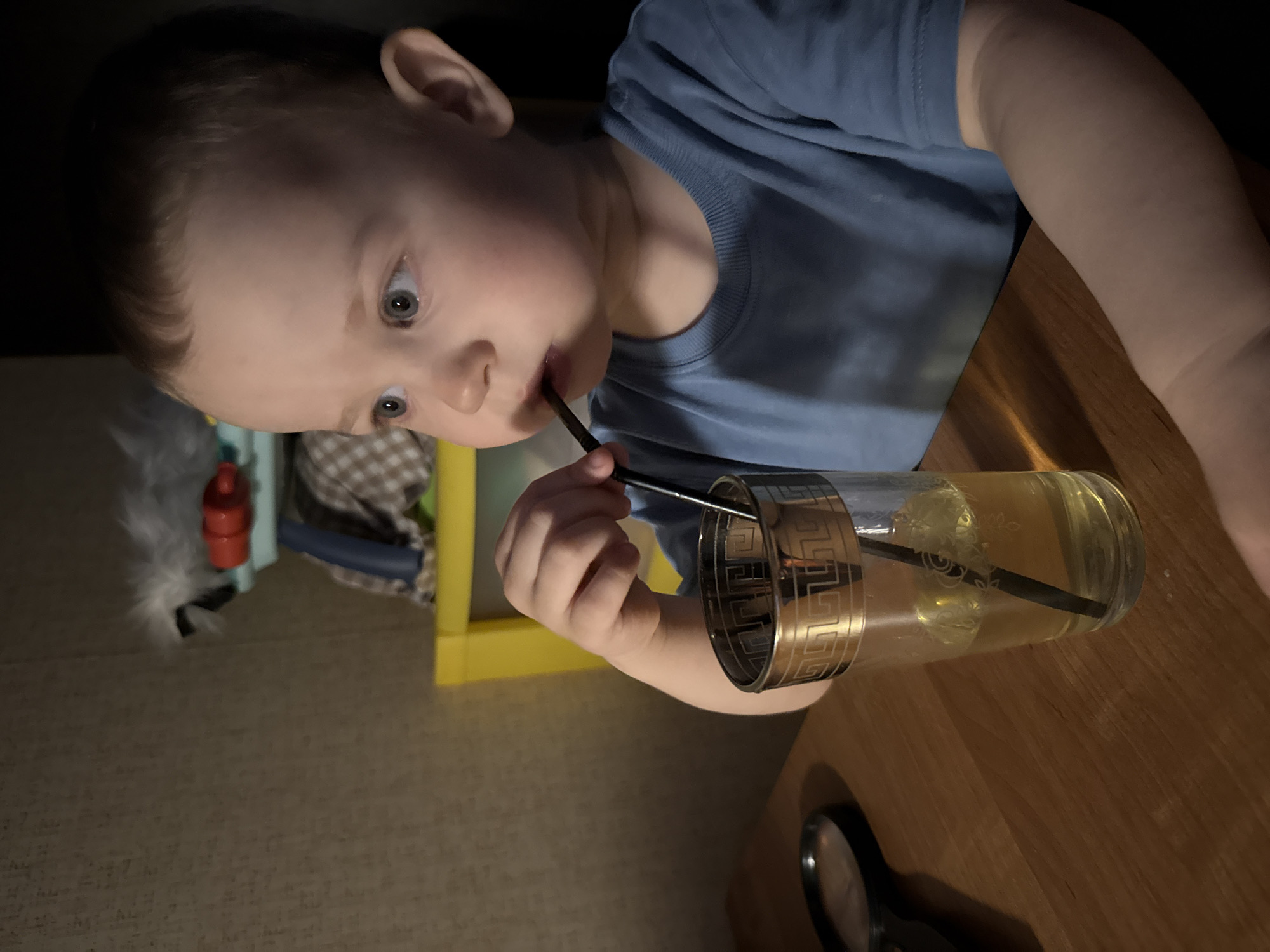 A young child, likely a toddler, is depicted indoors at a wooden table in Rusanovo, Russia. The child has light brown hair and wide, light blue eyes, and is wearing a long-sleeved blue top.
The primary activity shows the child holding a dark-colored straw to their mouth, appearing to be drinking from a tall, clear glass. The glass features a decorative metallic band with a Greek key pattern near its rim and contains a yellowish liquid with several ice cubes and the straw. The child's expression is one of focused concentration, looking slightly upwards.
In the foreground, partially visible on the wooden table, is a magnifying glass with a black handle. The background is dimly lit, suggesting an evening setting or artificial lighting. Hints of other household items are visible, including a yellow-framed structure, a red object, and a checkered fabric pattern, possibly a blanket or cushion. There is no discernible text visible in the image.