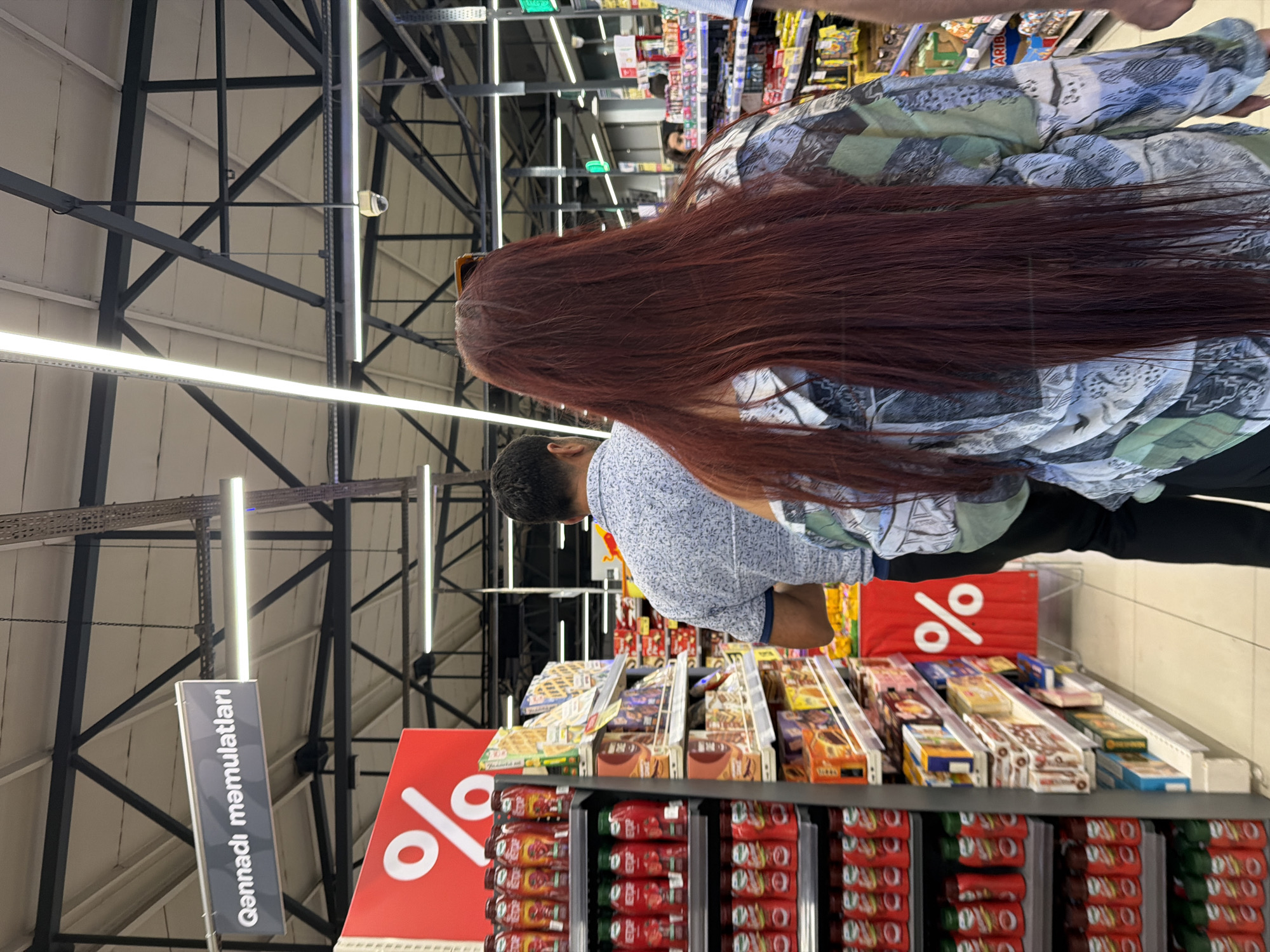 The image depicts the interior of a supermarket in Sumqayit, Azerbaijan, specifically within the "Qənnadı məmulatları" (confectionery) section. The scene is viewed from behind a woman with long, dark red hair, who is wearing a patterned shirt. In front of her, a man with short, dark hair is walking away from the camera, wearing a patterned shirt. To the left, shelves are stocked with numerous red bottles of what appear to be ketchup or similar sauces, many featuring the brand name "STROP." A large red sign with a white percentage symbol is also visible on the left. The ceiling features industrial lighting and structural beams, indicative of a large retail space. Other shoppers and shelves stocked with packaged goods are visible in the background, suggesting a busy shopping environment.