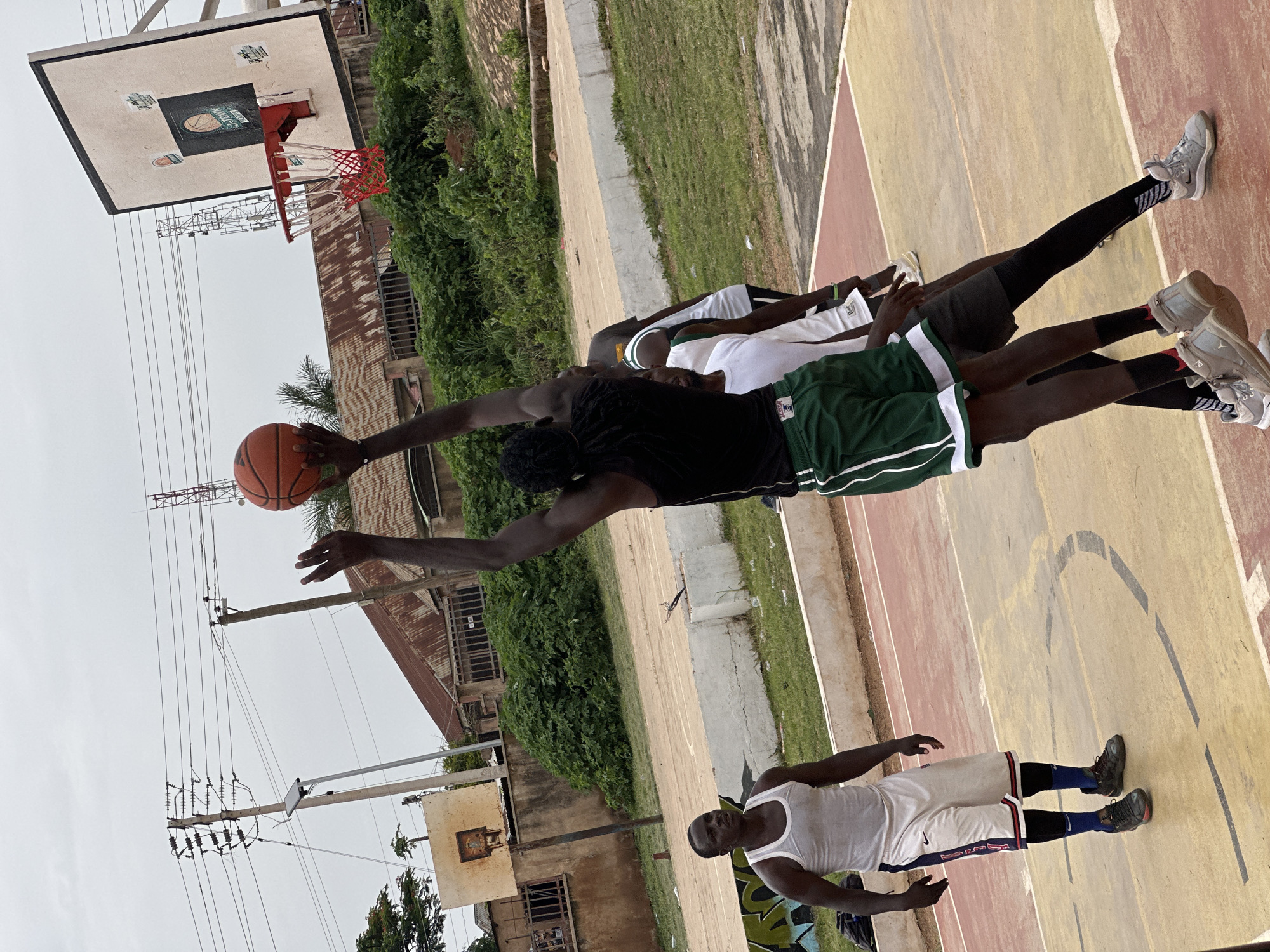 This image depicts an outdoor basketball game taking place on a worn court in Gura Top, Nigeria. Four Black men are visible on the court. The man in the foreground, on the left, wears a white tank top and white basketball shorts with red and blue trim, blue socks, and dark sneakers. He is facing forward and appears to be observing the game. Another man in the center of the frame, wearing a black sleeveless shirt and green basketball shorts with white trim, is in mid-air, reaching up to shoot a basketball. He is being defended by another man wearing a white t-shirt and dark shorts, who is partially obscured. A fourth man, wearing a dark t-shirt and dark shorts, is visible behind the man in white. The basketball court is made of asphalt with painted lines and has a concrete barrier along its edge. In the background, there are buildings with corrugated metal roofs, dense green vegetation, utility poles with overhead wires, and a basketball hoop. The sky is overcast and grey.