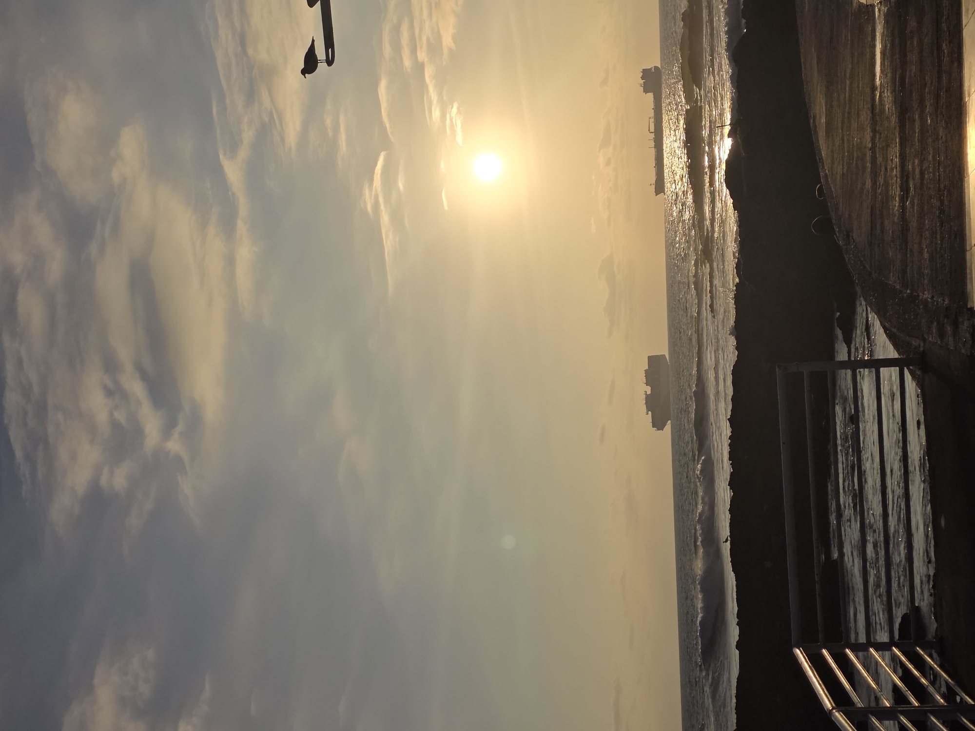 The image depicts a coastal scene at sunset in Iquique, Chile. The sun is low on the horizon, casting a warm golden light across the sky and sea, with scattered clouds tinged with pink and orange. Two large cargo ships are visible in the distance on the ocean. In the foreground, a rocky shoreline is partially visible, with waves crashing against it. A metal railing borders a walkway in the lower left of the frame. A seagull is perched on a lamp post in the upper right of the image. Another seagull is silhouetted on the rocks in the middle ground, facing the sea. The overall atmosphere is serene and peaceful, characteristic of a coastal sunset.