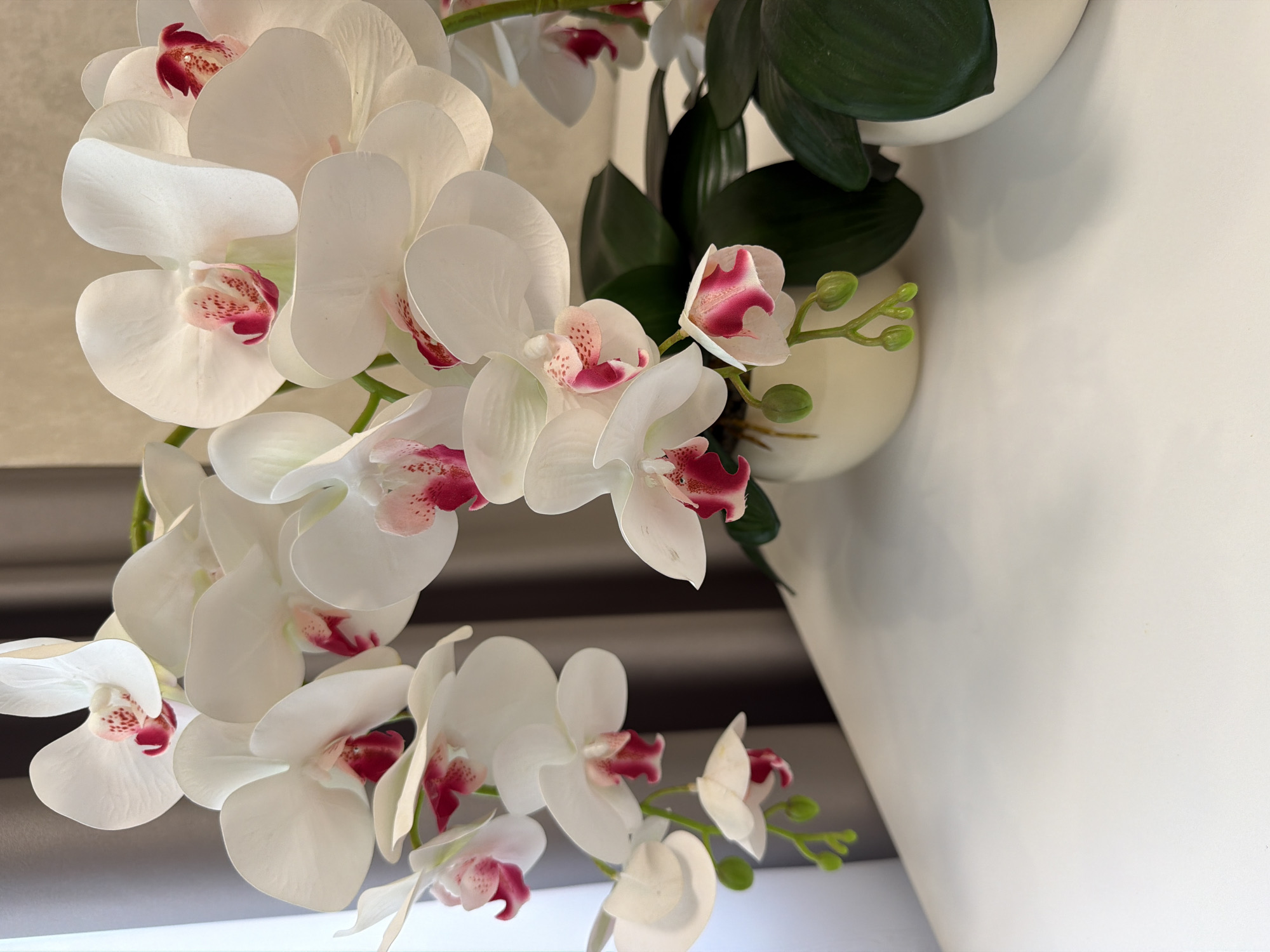 A close-up, eye-level shot focuses on a spray of artificial white orchids with pink centers, arranged in a cream-colored pot. The orchids are the primary subject, with their delicate petals and vibrant inner markings dominating the frame. Lush green leaves are visible towards the bottom right, peeking out from the pot. The background features a soft grey curtain, suggesting an indoor setting. The overall impression is one of elegant, artificial floral arrangement. The lighting is soft and diffused, casting gentle shadows.