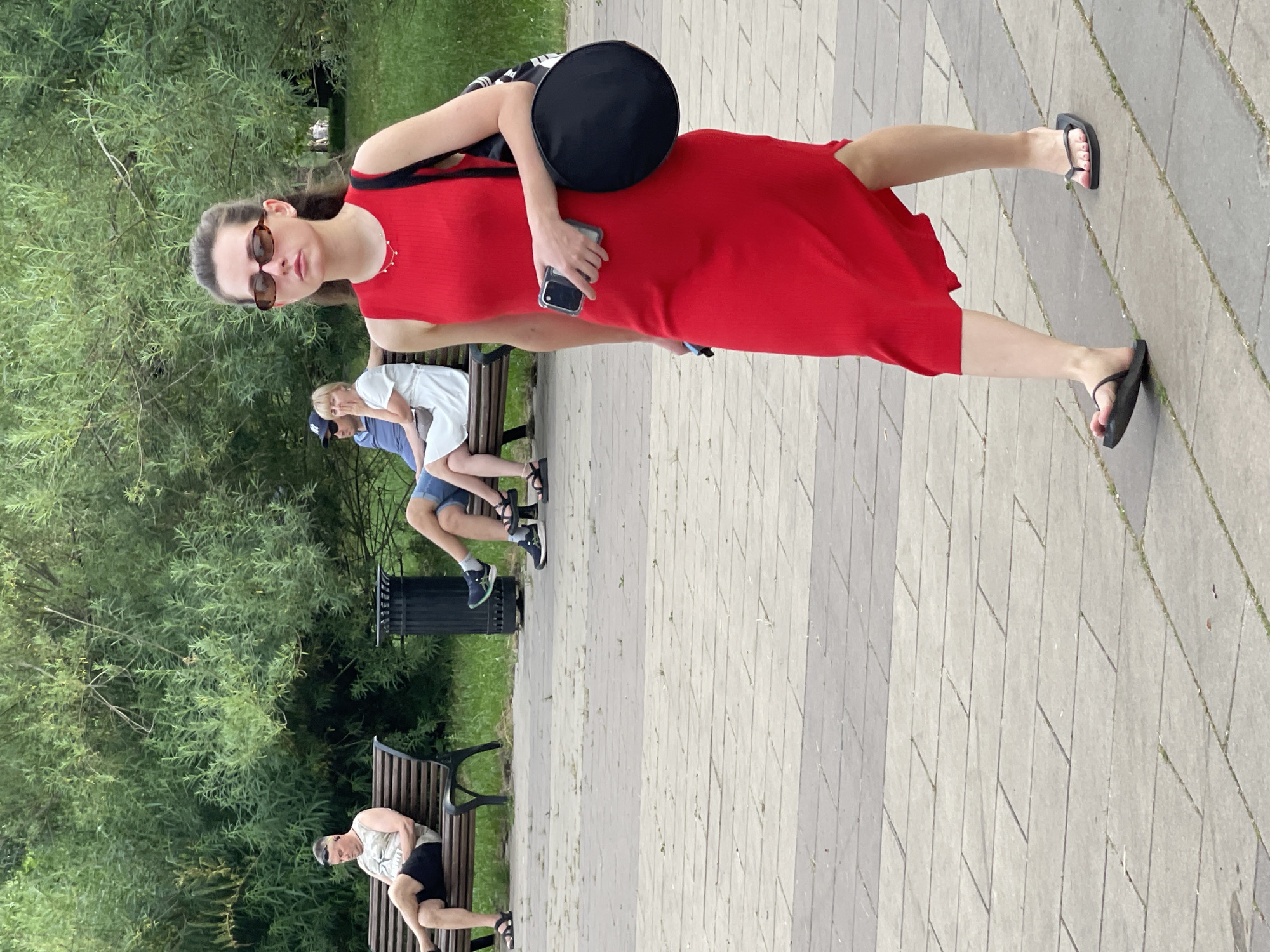 A woman in a red dress walks on a paved path in a park. She is wearing black flip-flops and sunglasses, and she is holding a smartphone in her right hand. Behind her, several people are sitting on park benches. The background consists of lush green trees and foliage. The scene appears to be a public park in Moscow, Russia, on a sunny day.