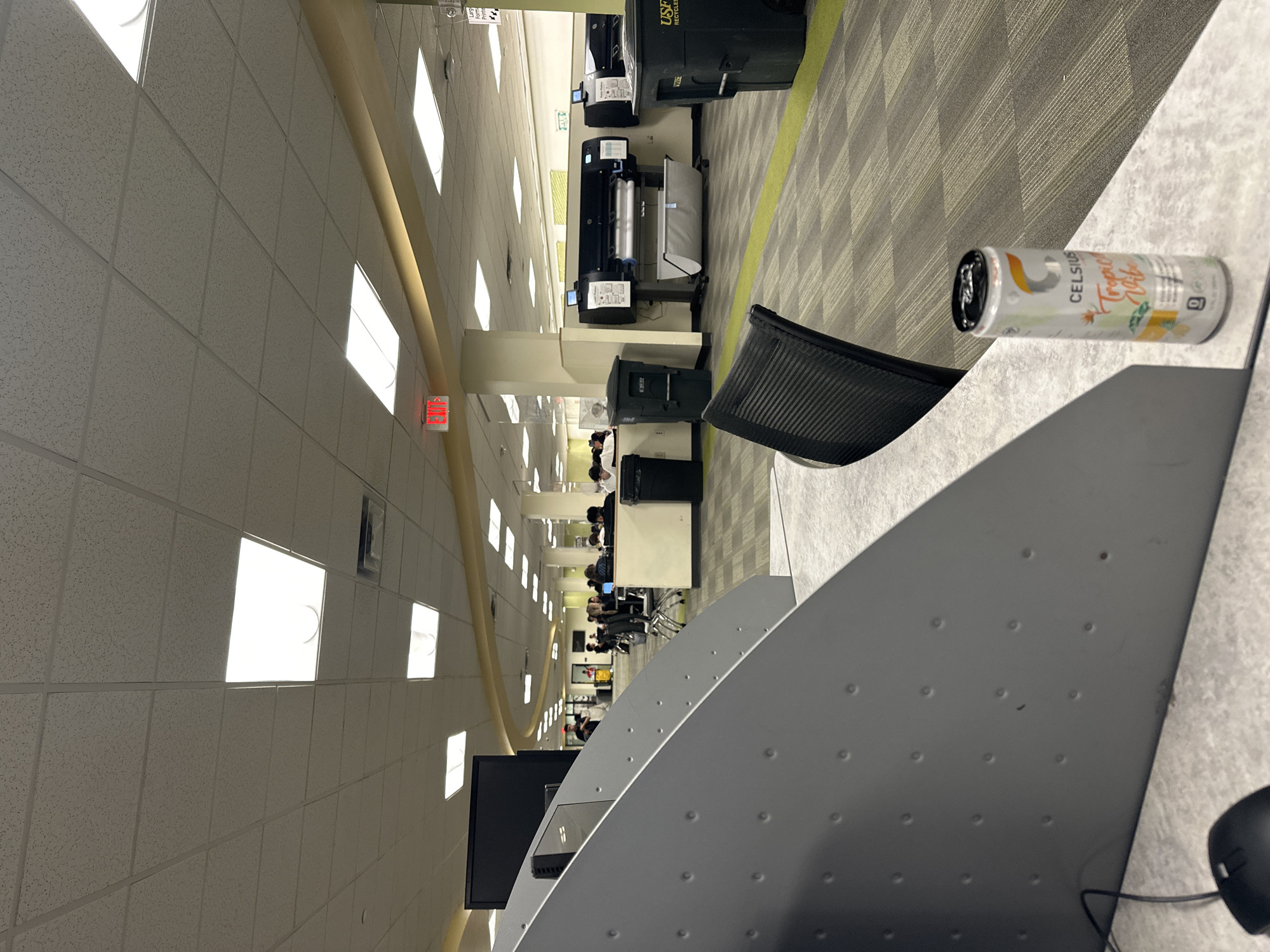 The image shows the interior of a library or study area, likely located in Tampa, United States of America. The view is slightly tilted, showcasing the rows of desks equipped with computers, suggesting a workspace for students or researchers. People are seated at some of the desks, presumably working or studying. Overhead, the ceiling is visible, fitted with recessed lighting panels arranged in a grid pattern. A large, beige-colored duct or architectural feature runs along the ceiling's length. There's an emergency exit sign visible in the distance. In the foreground, part of a workstation is visible, including a desk and a can of Celsius Tropical Vibe energy drink. A black office chair is also present. Further down the aisles, printers, trash receptacles, and other office equipment are located. The flooring consists of a patterned carpet.
