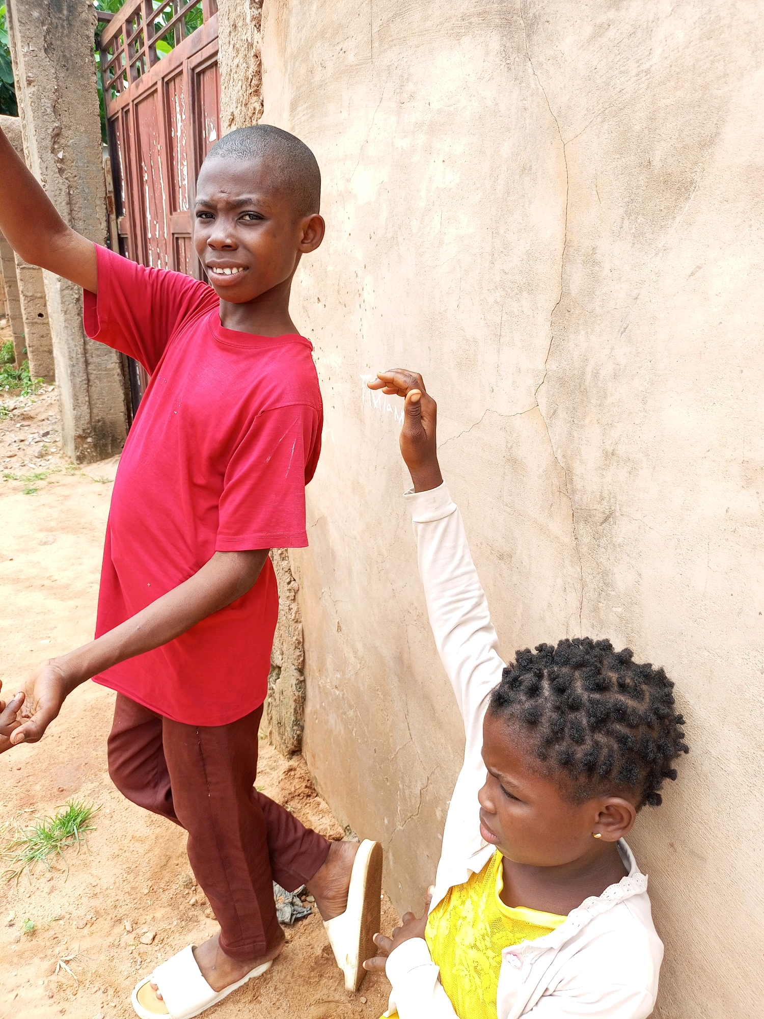 A young boy, dressed in a red t-shirt and brown pants, stands to the left of the frame, looking at the camera with a slightly apprehensive expression. His right arm is raised, and he appears to be holding something small and transparent in his hand. His left arm is extended downwards. He is wearing white slide sandals. To the right of the boy, a young girl is leaning against a textured, beige wall, looking to her left and upwards. She has short, dark hair styled in small twists and is wearing a yellow dress under a light-colored cardigan. Her right arm is raised, and her hand is near the wall. She is wearing white slide sandals as well. The ground beneath them is a sandy, dirt-like surface with sparse patches of green grass. In the background, a rusty metal gate is visible, partially obscured by a concrete pillar. The wall behind the children shows signs of wear and cracking. The overall setting appears to be an outdoor area in a residential neighborhood, likely in Tempure, Nigeria.