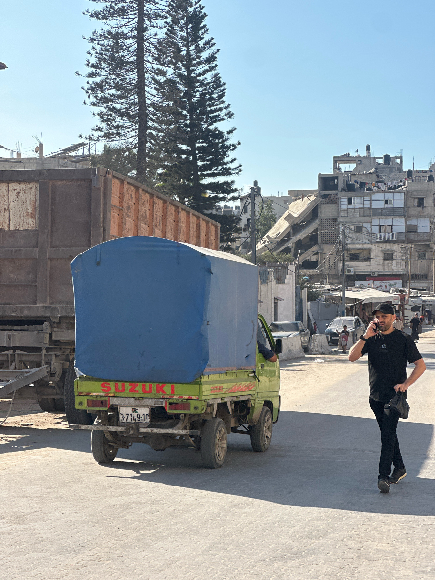 In the city of Gaza, Palestinian Territories, a man wearing a black t-shirt, black baseball cap, and black pants walks down a street while talking on his phone and holding a black bag. He is walking past a green Suzuki mini-truck with a blue tarp covering its bed. In the background, a large, partially destroyed building stands prominently, with debris scattered around its base. Other buildings, some appearing damaged or under construction, line the street. Two tall evergreen trees are visible on the left side of the frame, contrasting with the urban landscape. The sky is clear and blue. The street is paved with asphalt and appears to be a public road with several parked cars and some individuals in the distance.