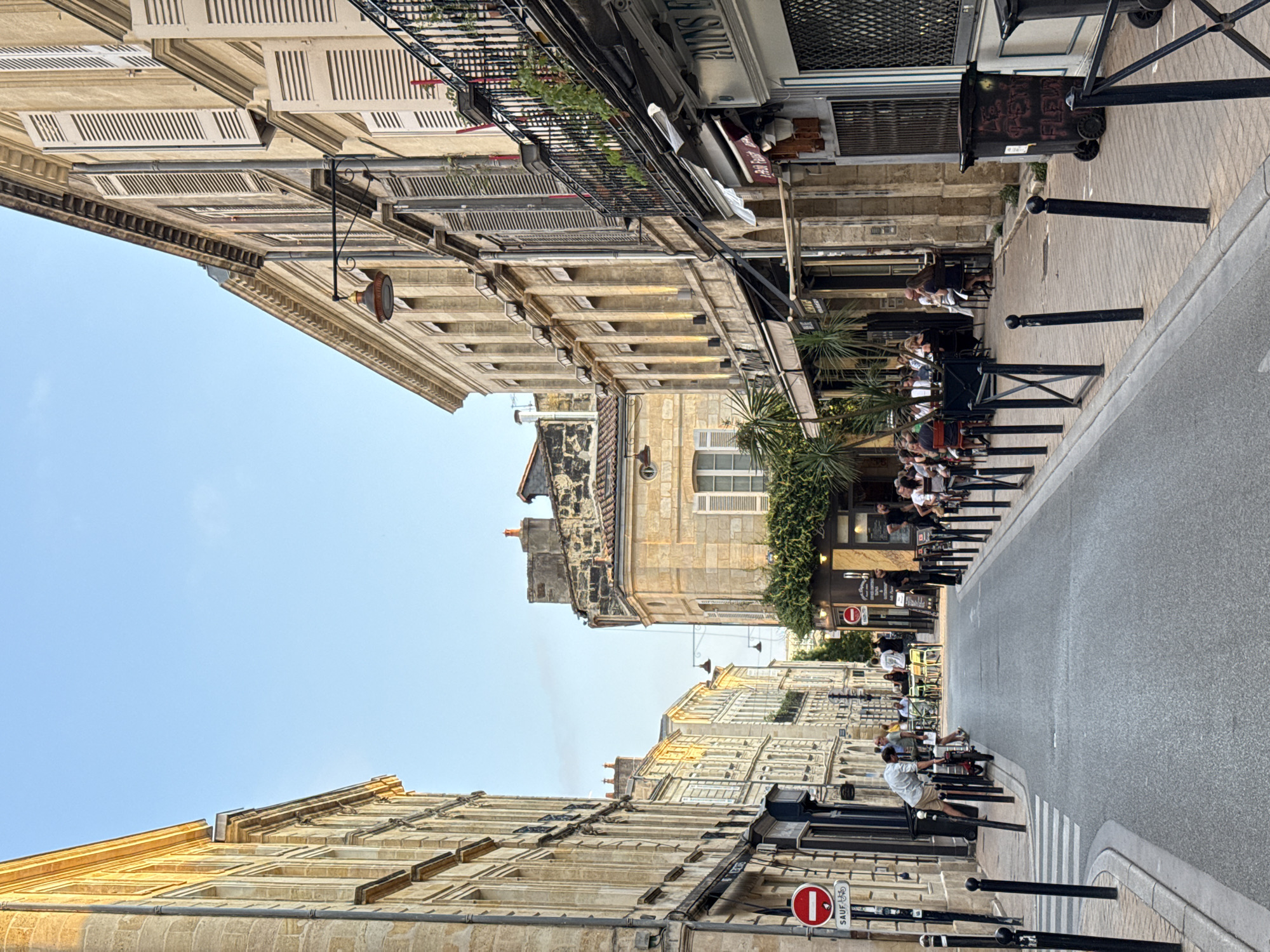 The image depicts a street scene in Bordeaux, France, likely in the historic city center, given the architecture. It is daytime, with clear blue skies and warm lighting on the stone buildings, suggesting it could be late afternoon or early evening. The street is paved and lined with traditional Haussmannian-style buildings constructed of light-colored stone, featuring arched windows, shutters, and ornate balconies.
On the left side of the street, several people are visible. A man in a light-colored shirt and dark pants is leaning over a red electric scooter near a pedestrian crossing and a "No Entry" sign. Further down, a group of people are seated at tables outside a building, some appearing to be dining or socializing.
The right side of the street is also lined with buildings, housing businesses that appear to be cafes or restaurants, indicated by awnings, outdoor seating, and people gathered at tables. One establishment has a sign that is partially visible and reads "PAINS F..." suggesting a bakery or food service. A large black trash bin with some graffiti is positioned on the sidewalk.
The street is bordered by black bollards and a paved sidewalk. The overall impression is that of a lively, urban environment with a blend of residential and commercial activity, typical of European cities.