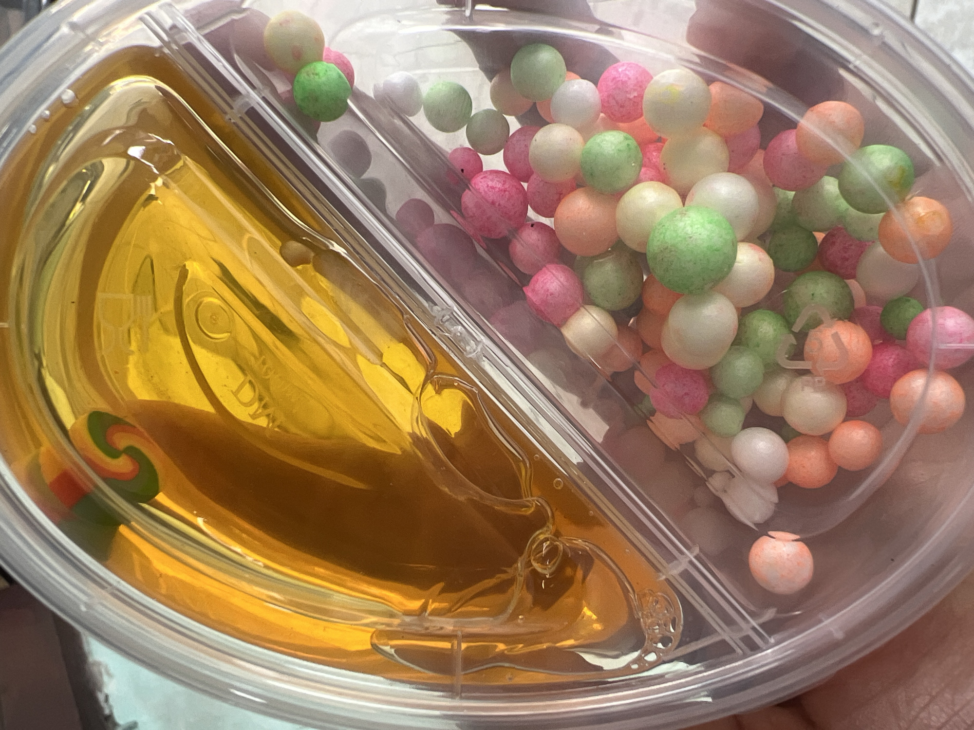 A transparent plastic container is filled with two different substances. The left compartment contains a viscous, golden-yellow liquid, possibly honey or a similar syrup, with a small swirl-patterned object partially submerged within it. The right compartment is filled with a multitude of small, round, colorful beads in shades of pink, green, orange, and white, resembling styrofoam balls or candy. The container is held in a person's hand, suggesting it is a personal item or product. The background is blurred, but hints of a light blue surface and shadows are visible. The image was taken in Gunung Putri, Indonesia.