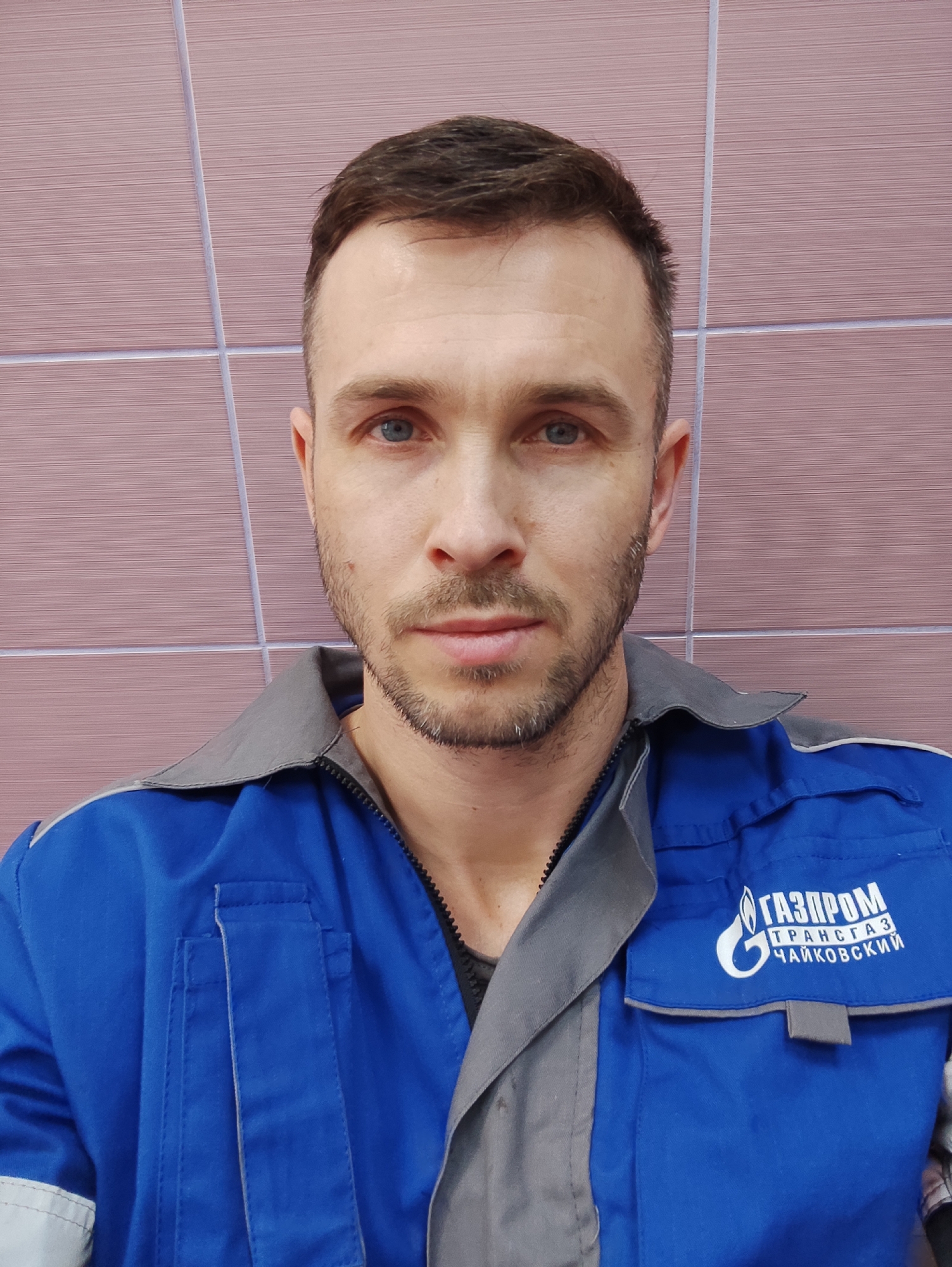A man is wearing a blue uniform with a gray collar and the logo of "GAZPROM TRANSGAZ CHAIKOVSKIY" on the right breast. He has short brown hair, a beard, and blue eyes. He is indoors in front of a tiled wall. The city is Unknown, Unknown.