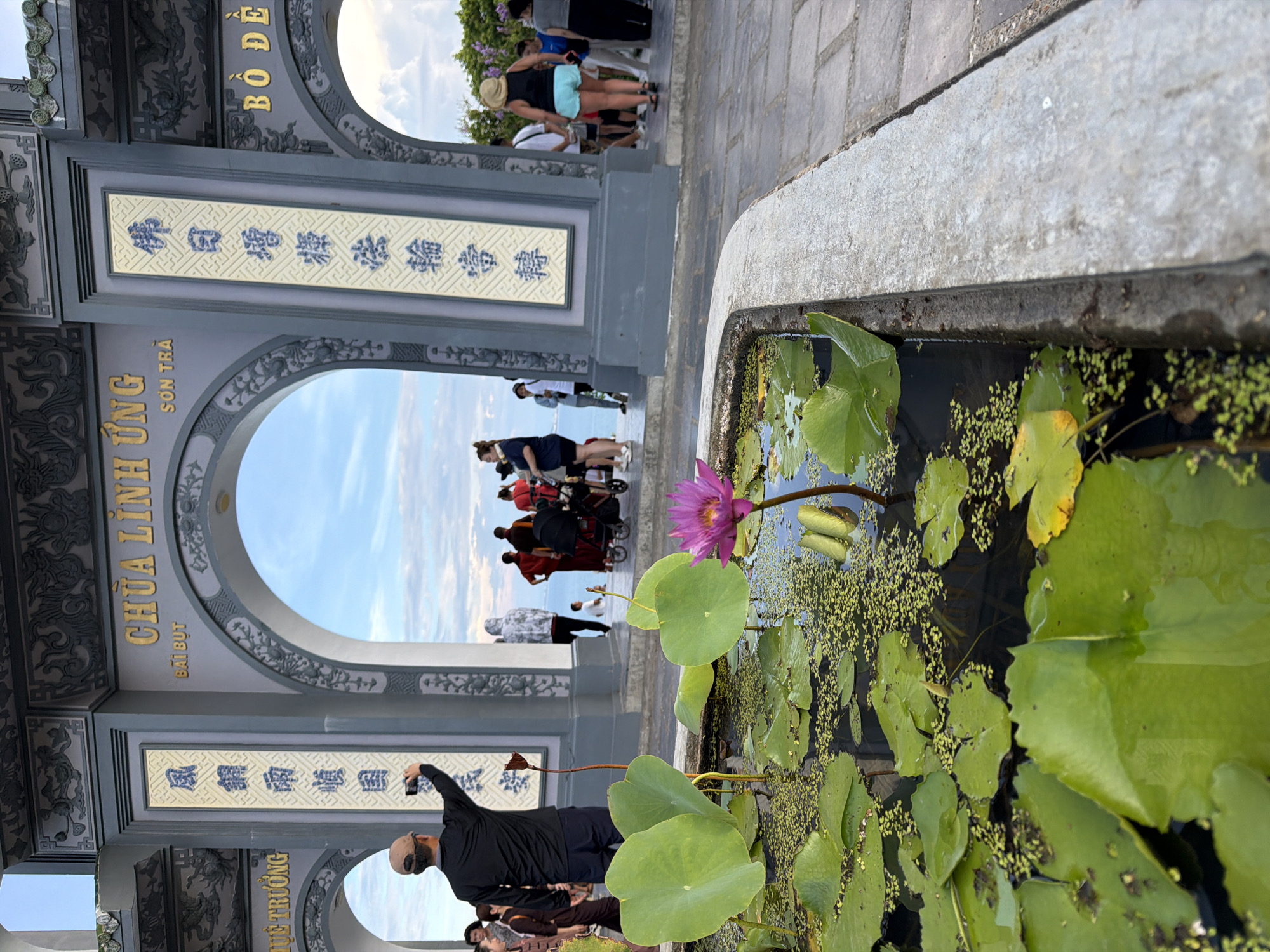 The image captures a view through a grand temple gate with arched openings, revealing a scenic backdrop of the ocean and sky. In the foreground, a lotus pond with vibrant green lily pads and a single pink lotus flower is prominently featured. A man wearing a dark shirt and a beige cap is seen from behind, holding up a mobile phone to take a picture. Behind him, through the central arch, several people are visible, some dressed in traditional Vietnamese attire, looking out towards the sea. Two children are also present, one near a stroller. The temple gate itself is adorned with intricate carvings and vertical inscriptions in both Vietnamese and Chinese characters. The overall scene suggests a visit to a cultural or religious site in Đà Nẵng, Vietnam, likely the Linh Ứng Pagoda on Sơn Trà Peninsula, known for its stunning ocean views and large Buddhist statues. The lighting indicates it is daytime, possibly late afternoon, with soft sunlight illuminating the scene.