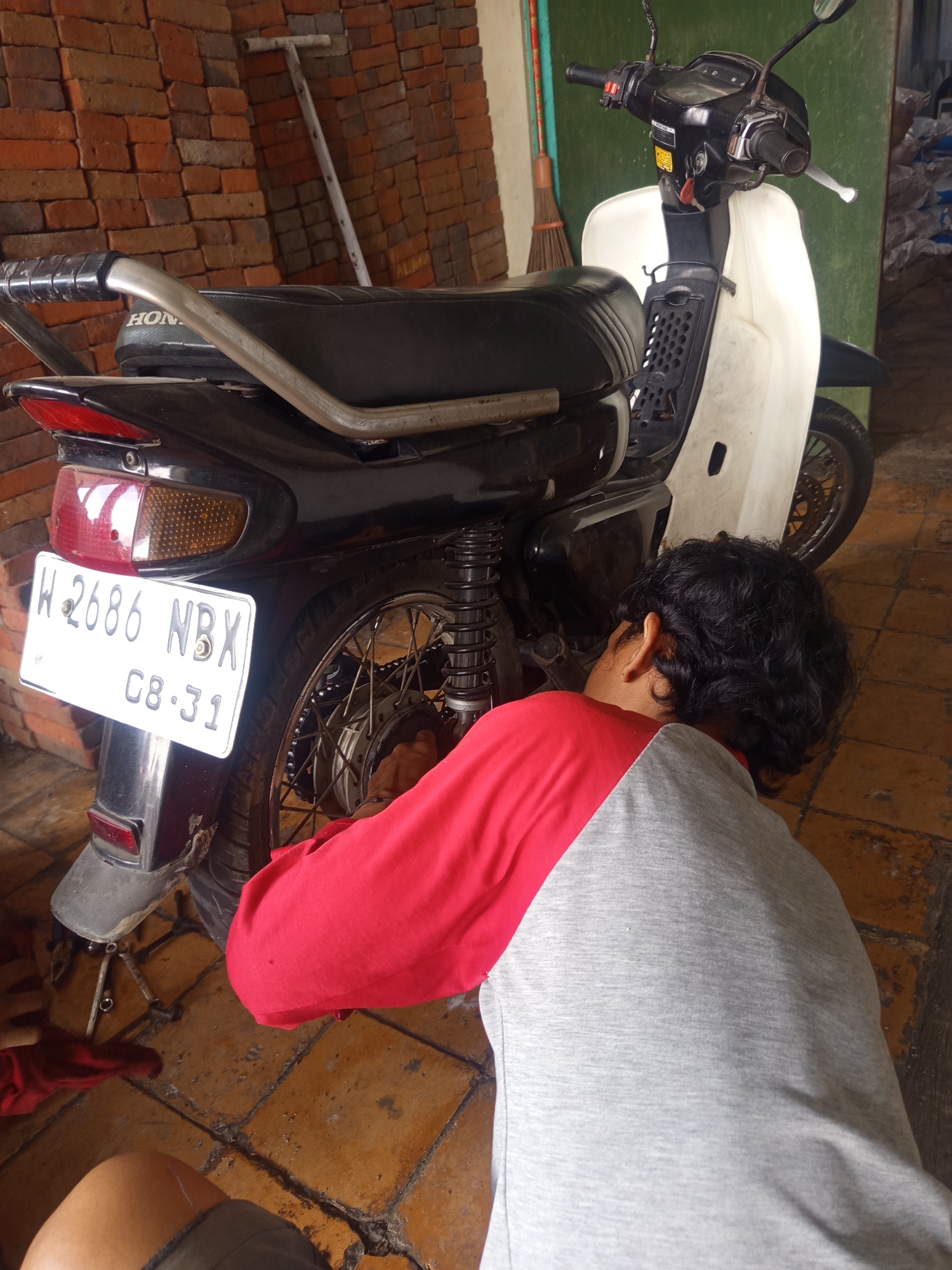 A person is working on the rear wheel of a black and white Honda motorcycle, which is parked on a tiled floor indoors. The motorcycle has a license plate that reads "W 2686 NBX 08.31". The person is wearing a red and grey shirt and has dark curly hair. Behind the motorcycle, there is a wall made of red bricks and a broom leaning against it. The location is the city of Sumber Gedang, Indonesia.